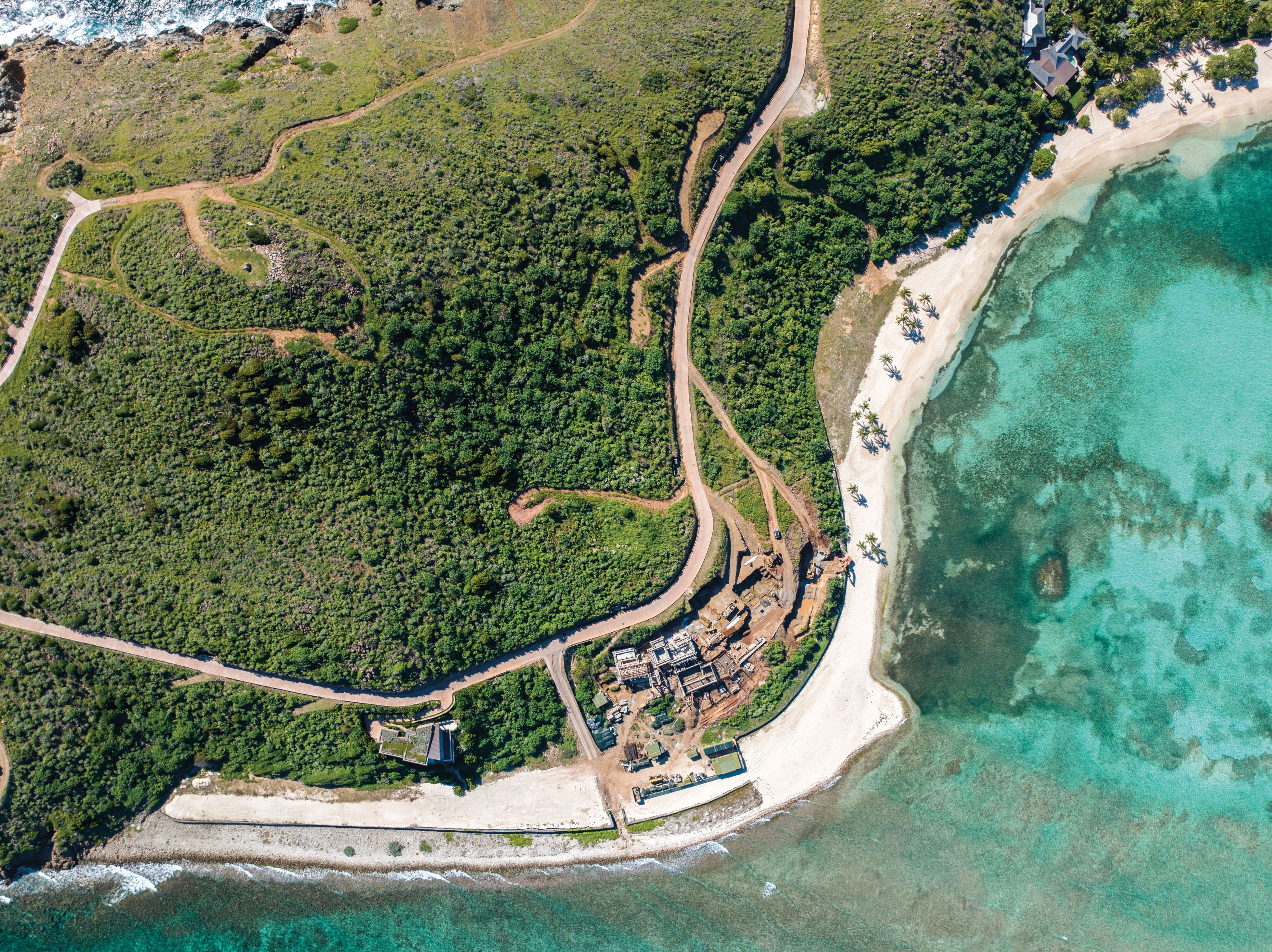 Aerial view of a peninsula coastline with a green hillside featuring winding dirt paths, and partially constructed or ruined stone structures near the shore. The shoreline has a sandy beach merging with clear turquoise waters. Forested areas surround the construction.