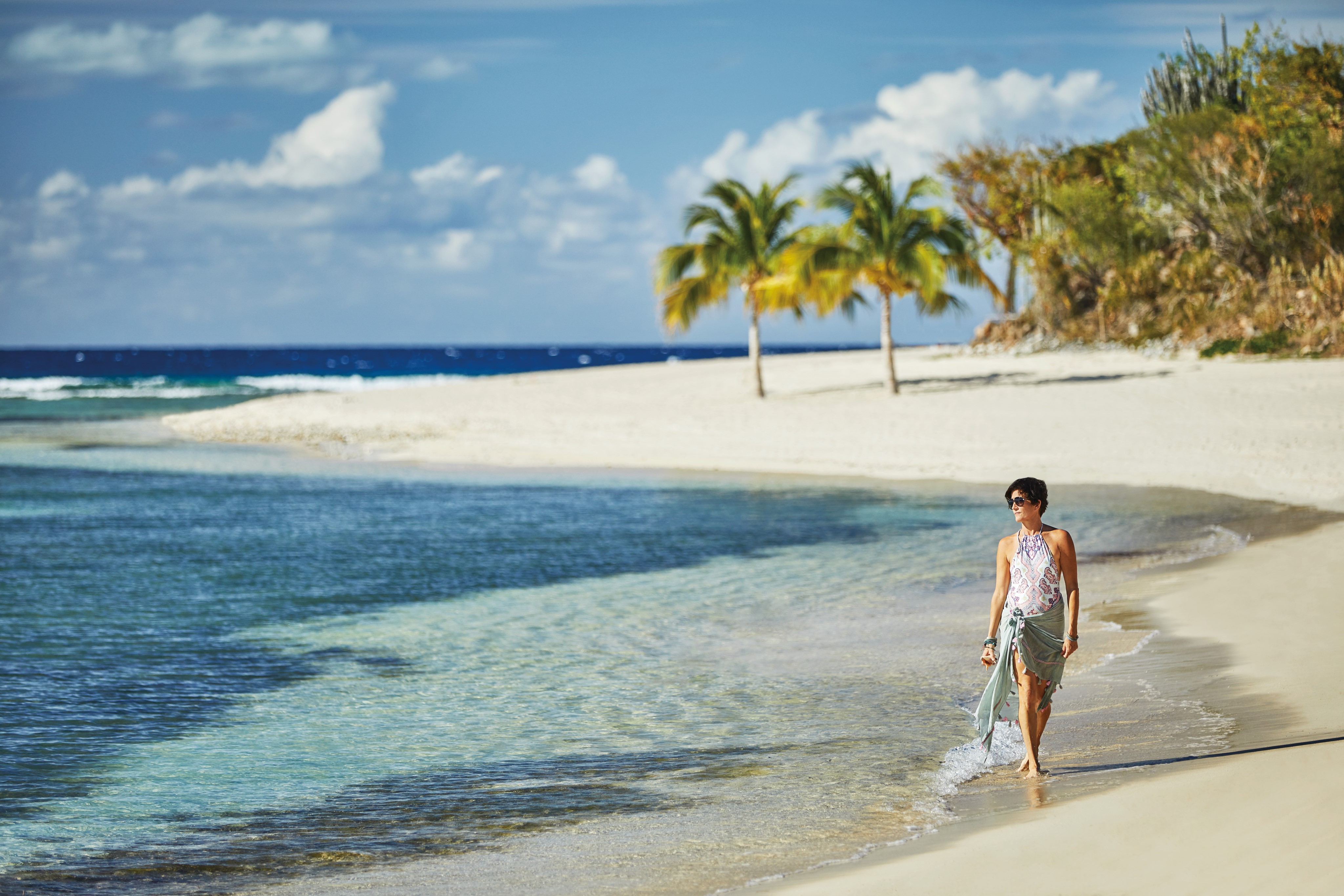 A person in casual beachwear walks along the shoreline of a tropical beach. The clear blue water meets the white sandy shore of the peninsula, with a backdrop of palm trees and a partly cloudy sky.