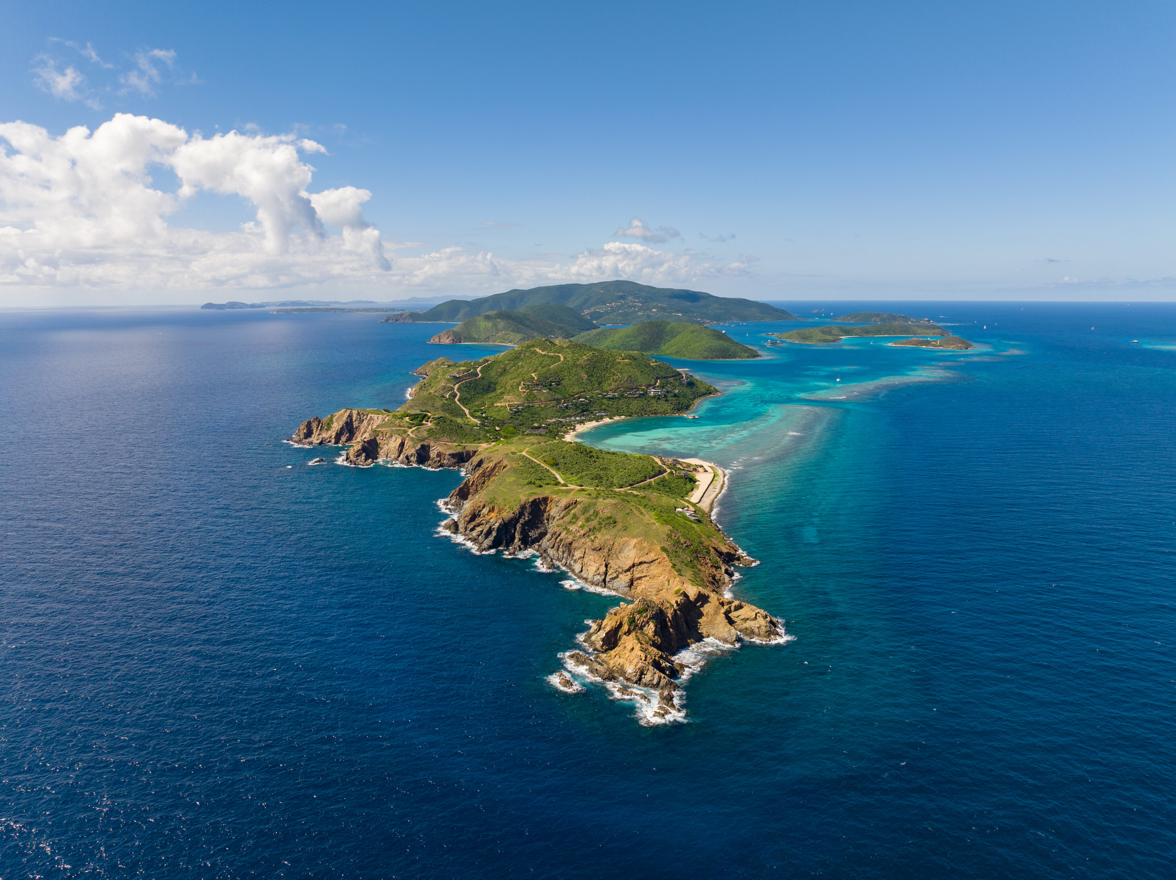 Aerial view of a long, narrow island surrounded by the clear blue ocean waters of Pajaros Point. The island features rocky coastlines, lush green terrain, and white sandy beaches. The sky is clear with a few scattered clouds. Smaller islands are visible on the horizon.