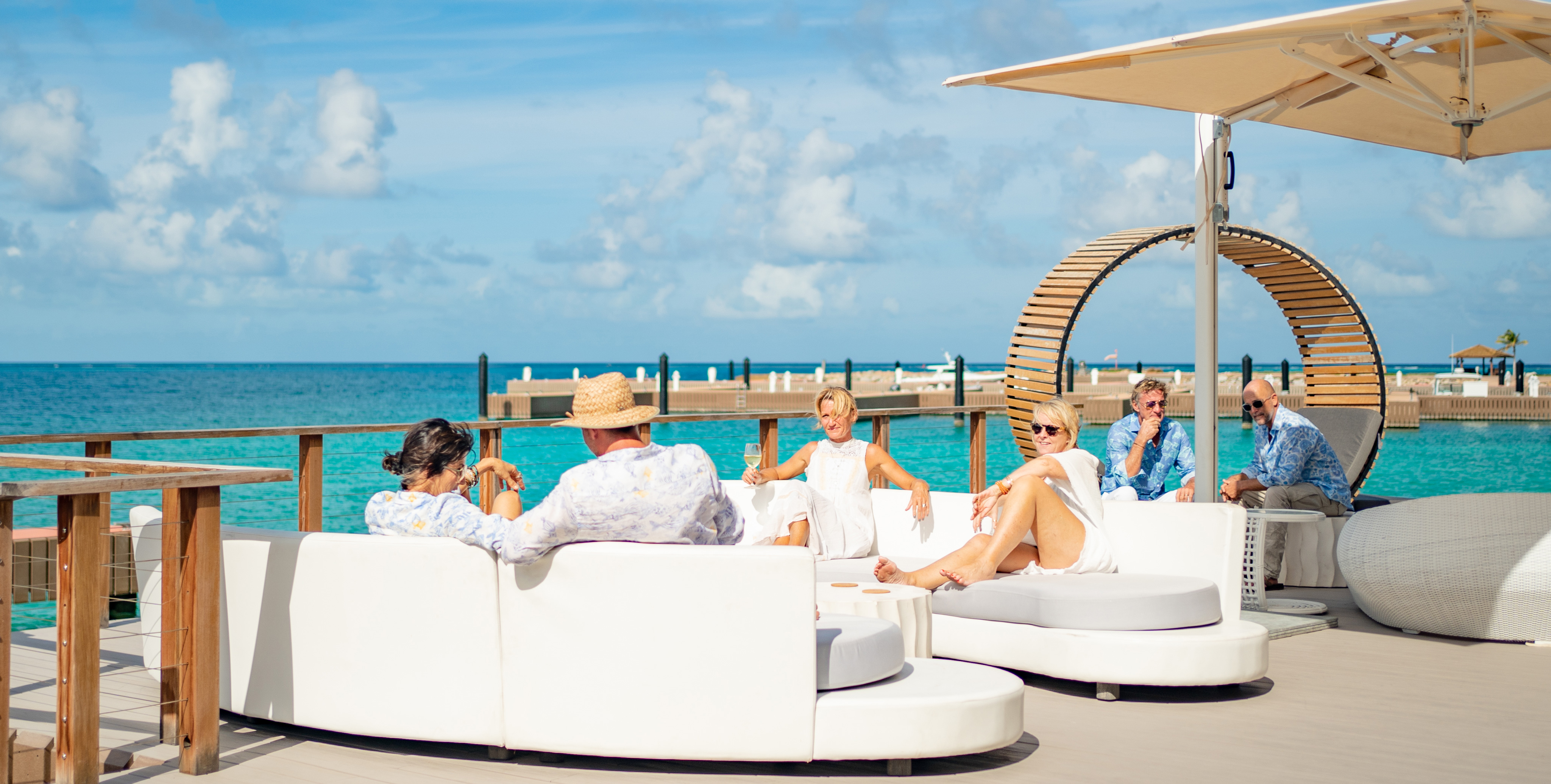 A group of people is relaxing on white couches on a sunny deck by the sea. They are engaged in conversation under a large Nova umbrella, with a circular wooden structure and clear turquoise water in the background.