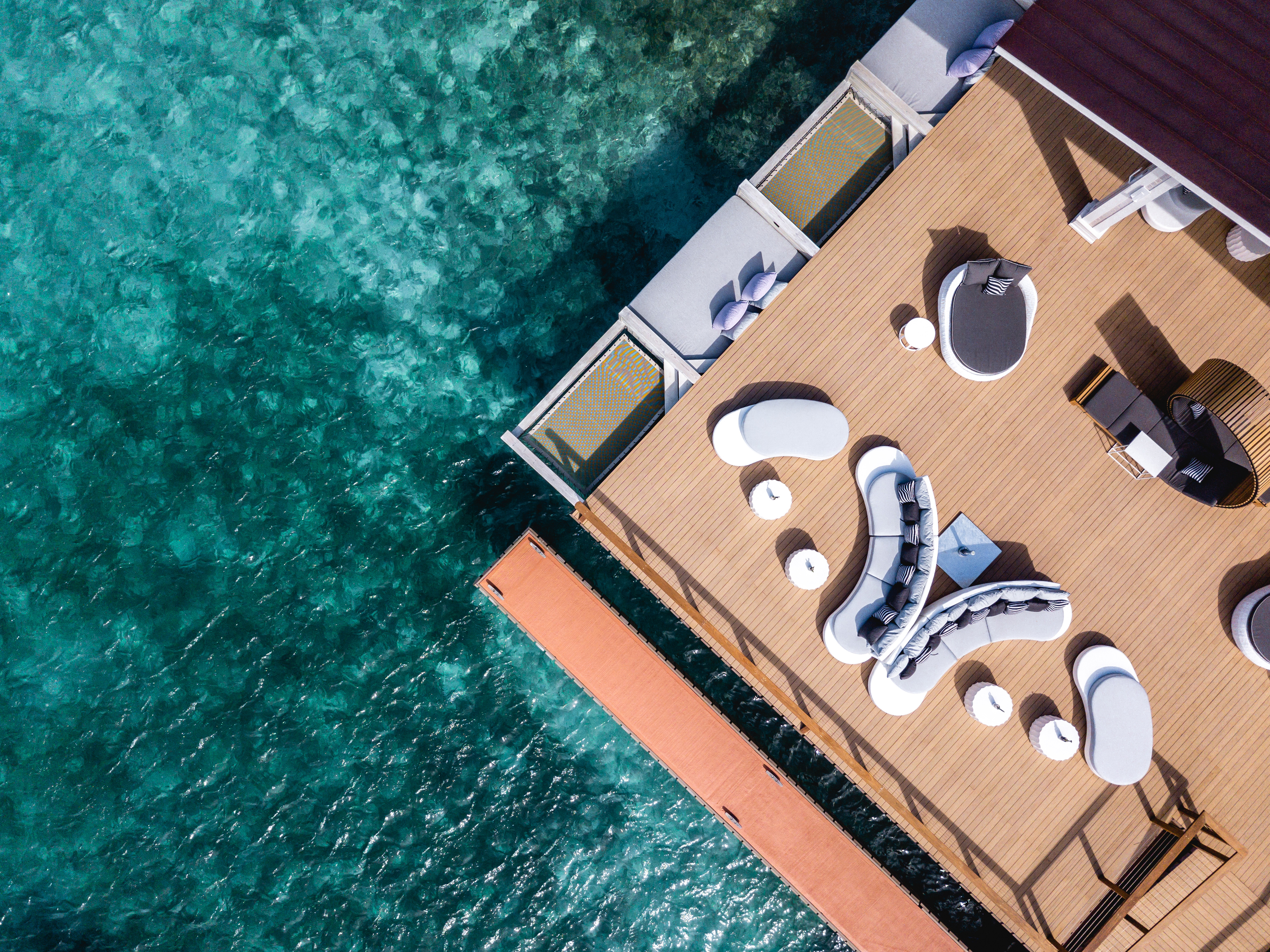 Aerial view of Nova's wooden deck over a turquoise ocean. The deck features modern white and gray lounge furniture arranged in amorphous shapes. A narrow red-brown walkway extends from the deck over the water, inviting you to explore further into this serene paradise.