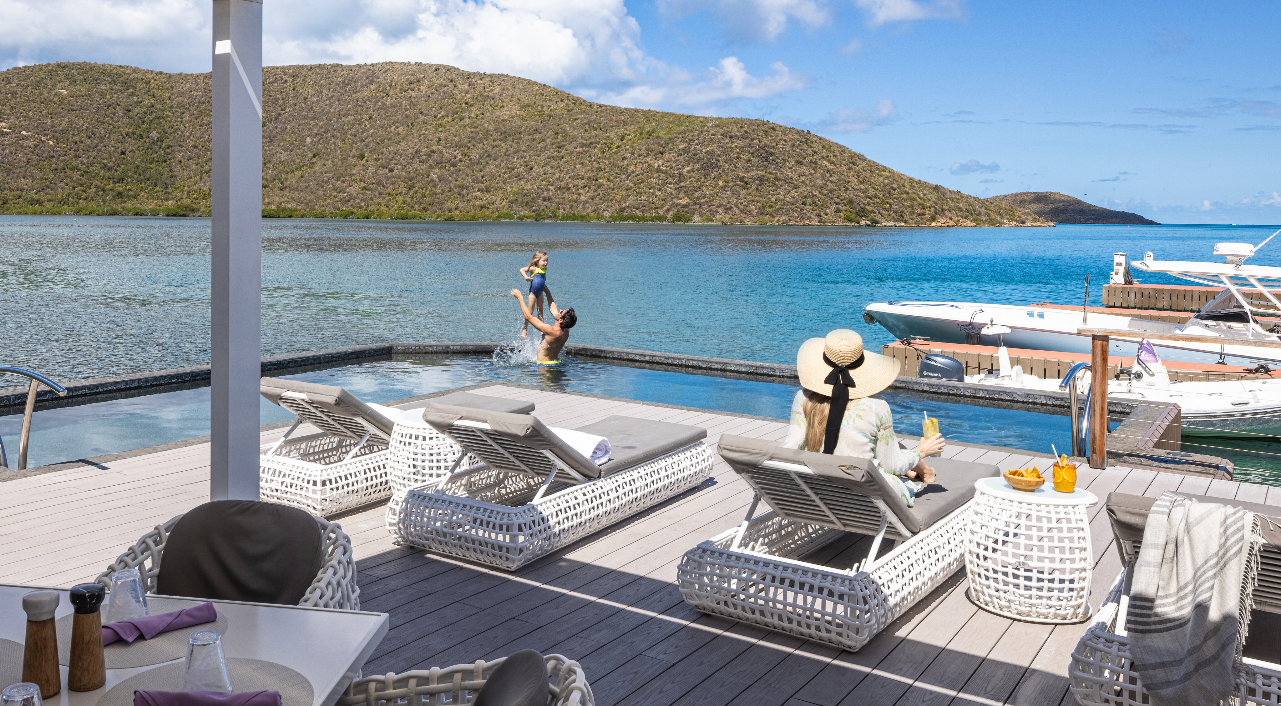 A serene seaside scene featuring two people lounging on white poolside chairs under a shaded canopy, with a person in the water lifting a child. Boats named "Nova" are docked nearby, and distant hills are silhouetted against a blue sky with clouds.