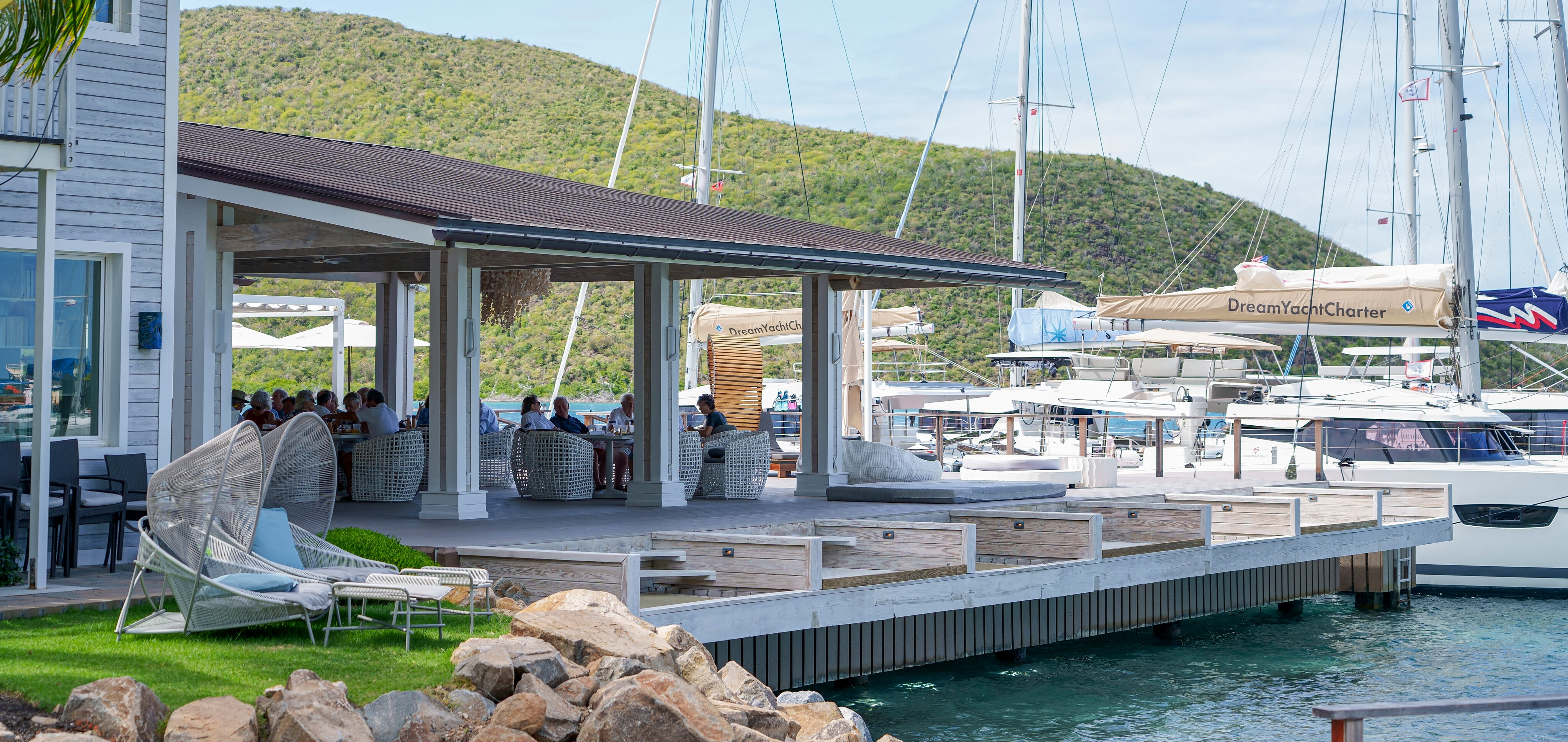 The Nova marina boasts several sailboats docked under the sun, with a wooden deck offering seating for those relaxing beside a charming building. In the backdrop, lush green hills rise beneath a partly cloudy sky.