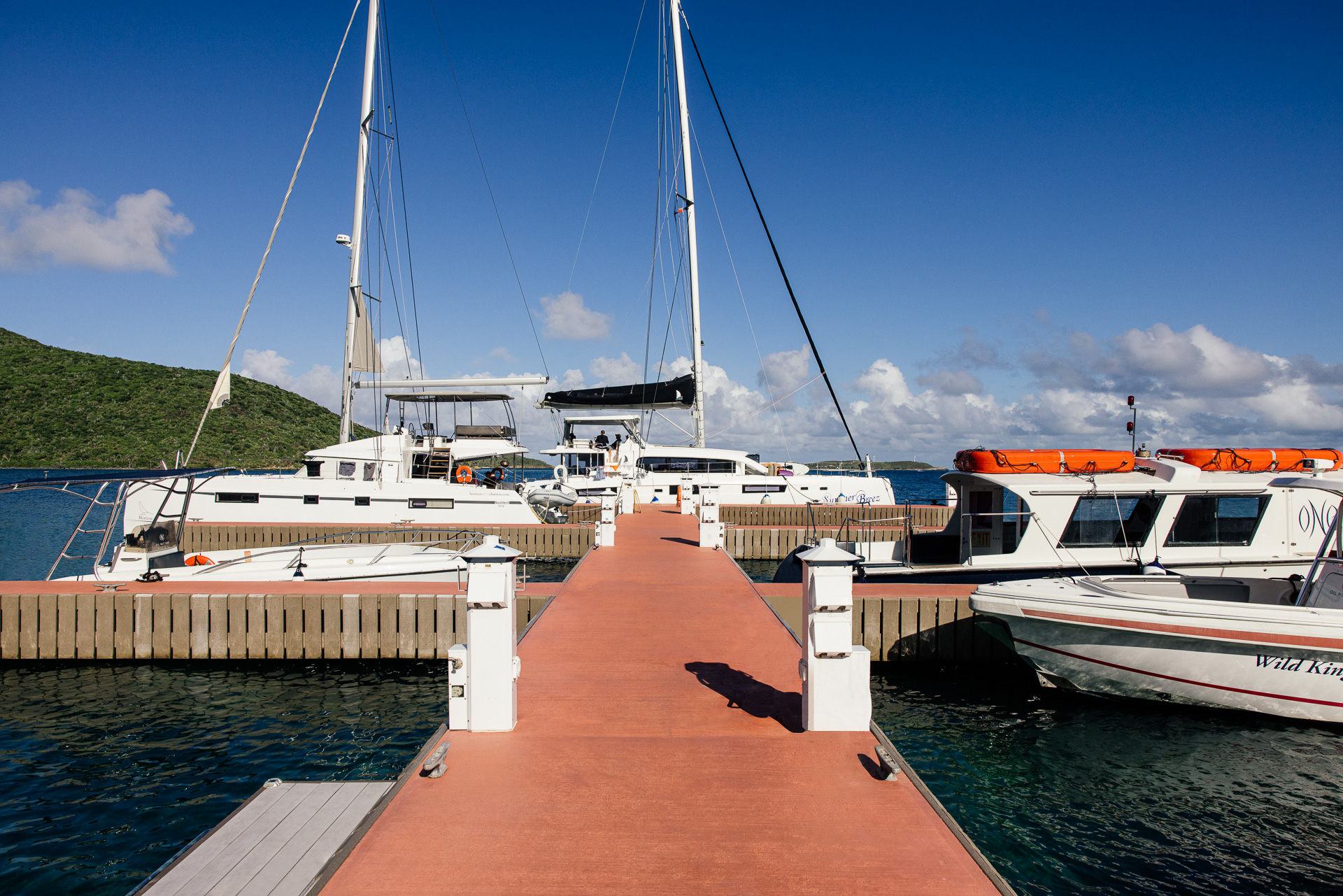 A wooden pier extends into Nova Marina with multiple docked sailboats and motorboats on a calm, blue sea. The boats have white hulls and canopies, and the sky is clear with a few scattered clouds. In the background, there is a green hillside to the left.