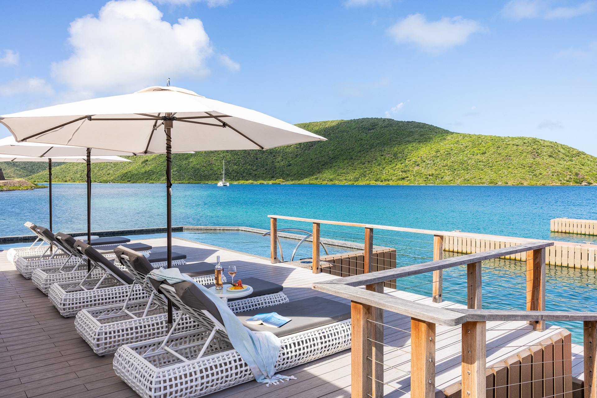 A serene view of a coastal deck featuring cushioned lounge chairs shaded by umbrellas facing a vibrant blue Nova ocean. The deck overlooks a lush green hillside across the water, and a small boat can be seen in the distance under a partly cloudy sky.