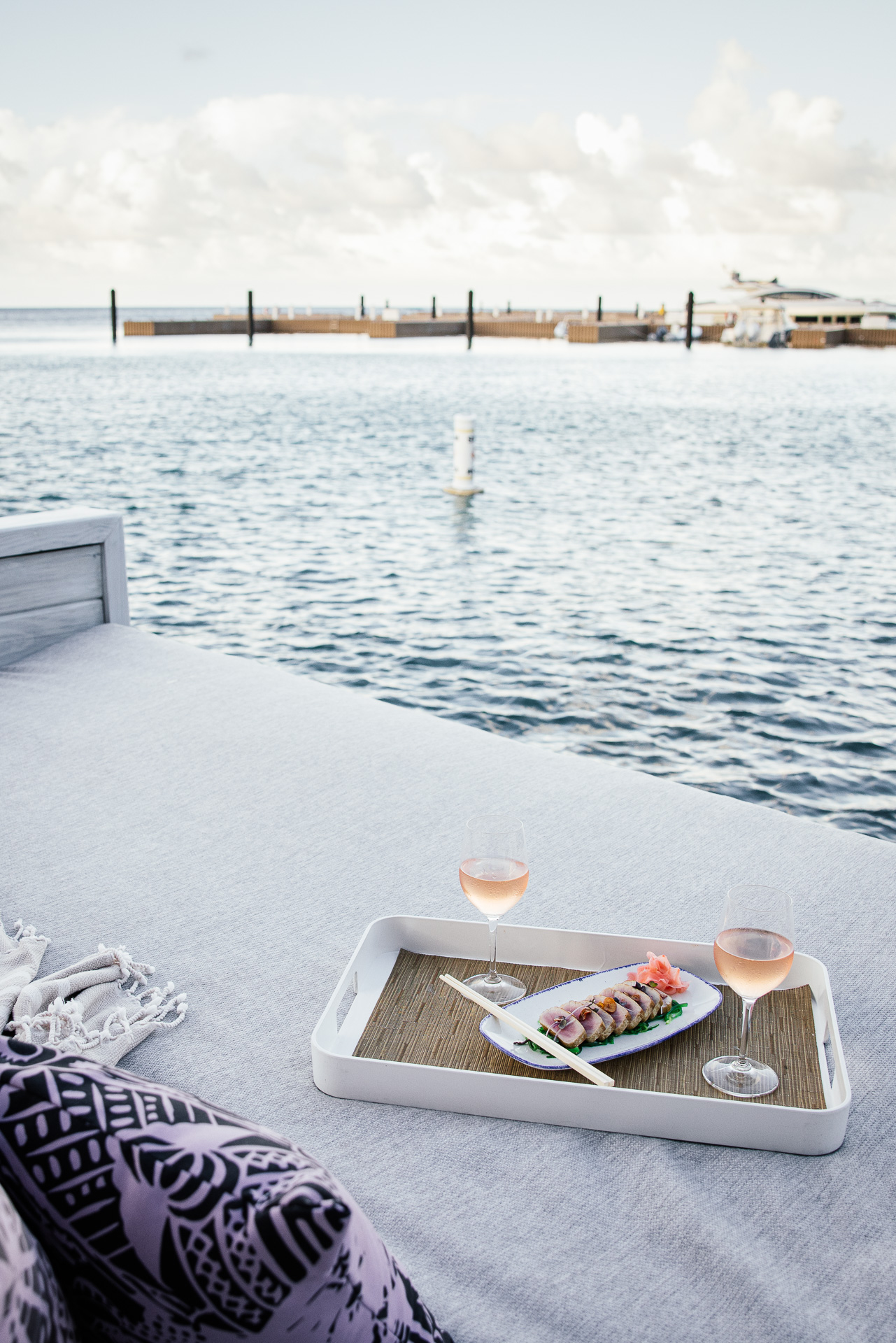 A serene waterside view featuring a tray with sushi and two glasses of rosé wine on a Nova platform bed. The bed overlooks a calm body of water with docks and a cloudy sky in the background. Cushions and a light blanket add to the cozy setting.