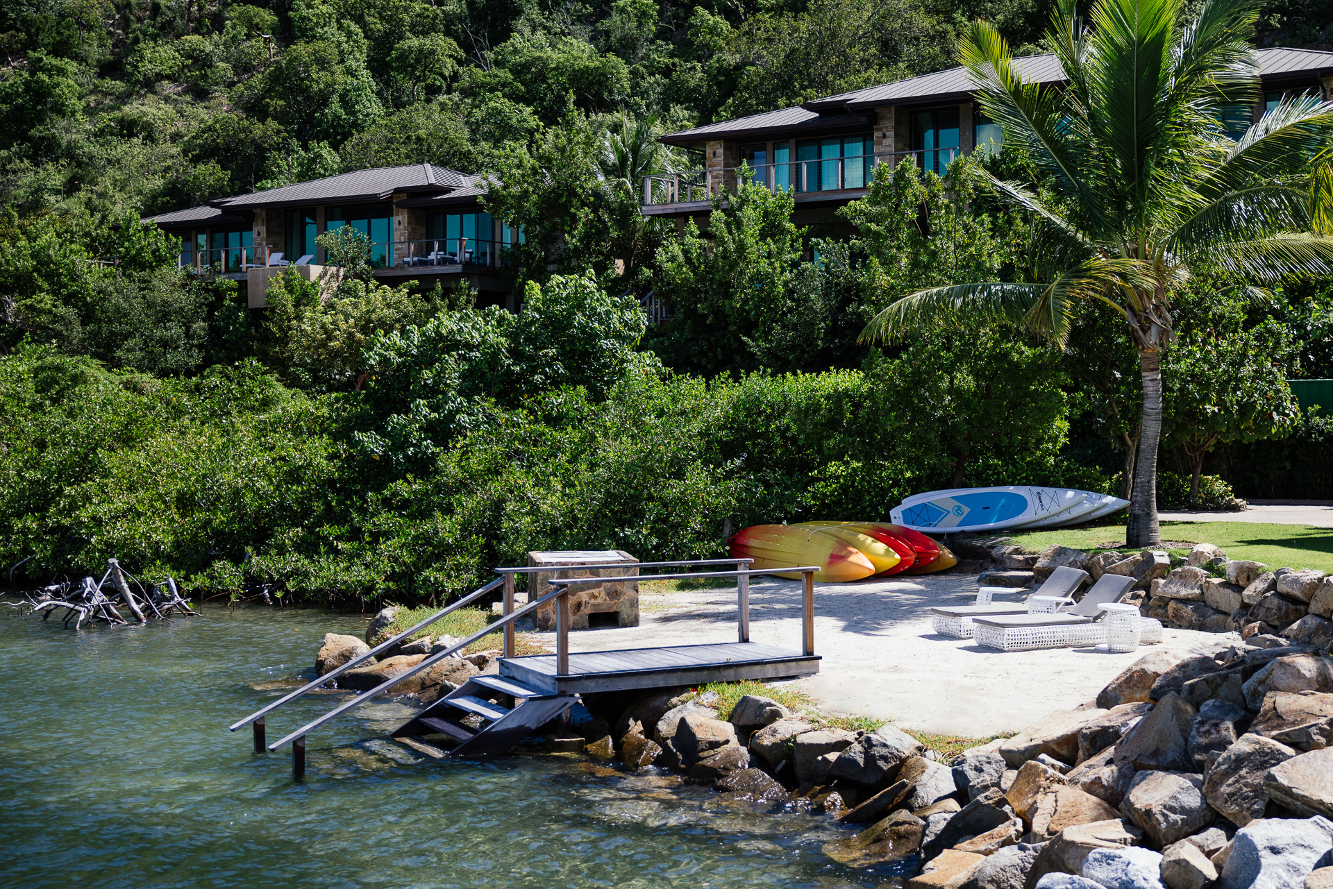 A serene beachfront setup with a small dock, Nova kayaks, and paddleboards resting on a sandy area. In the background, lush green foliage surrounds elegant villas perched on a slight hill. A palm tree adds to the tropical ambiance of the scene.