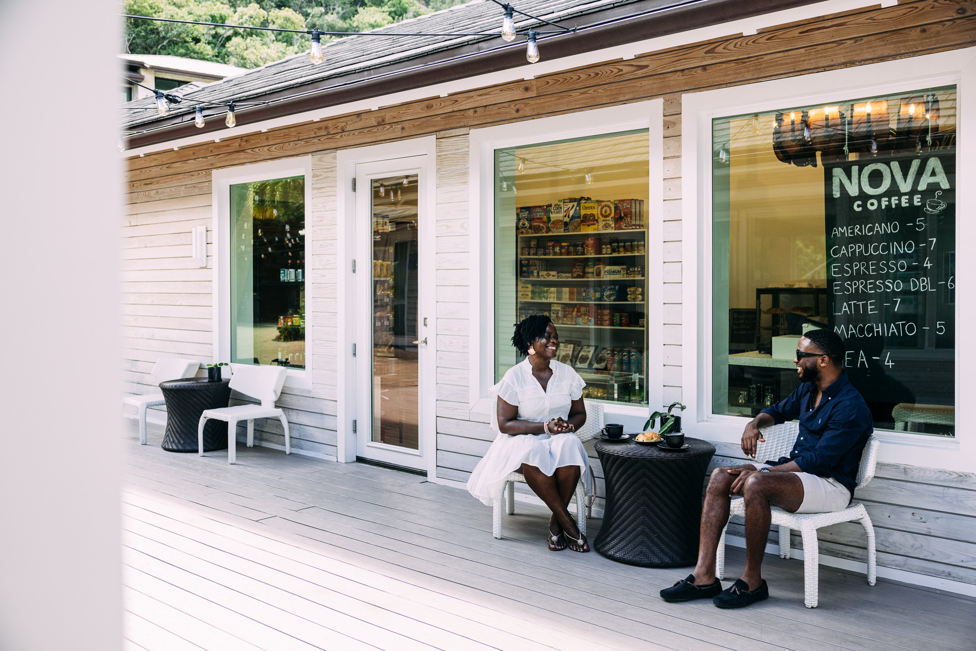 Two people sit outside Nova, a coffee shop with a rustic wooden exterior. They are seated at a small round table with drinks. String lights hang above them, and a large menu is visible on the window behind them. The mood is relaxed and casual.