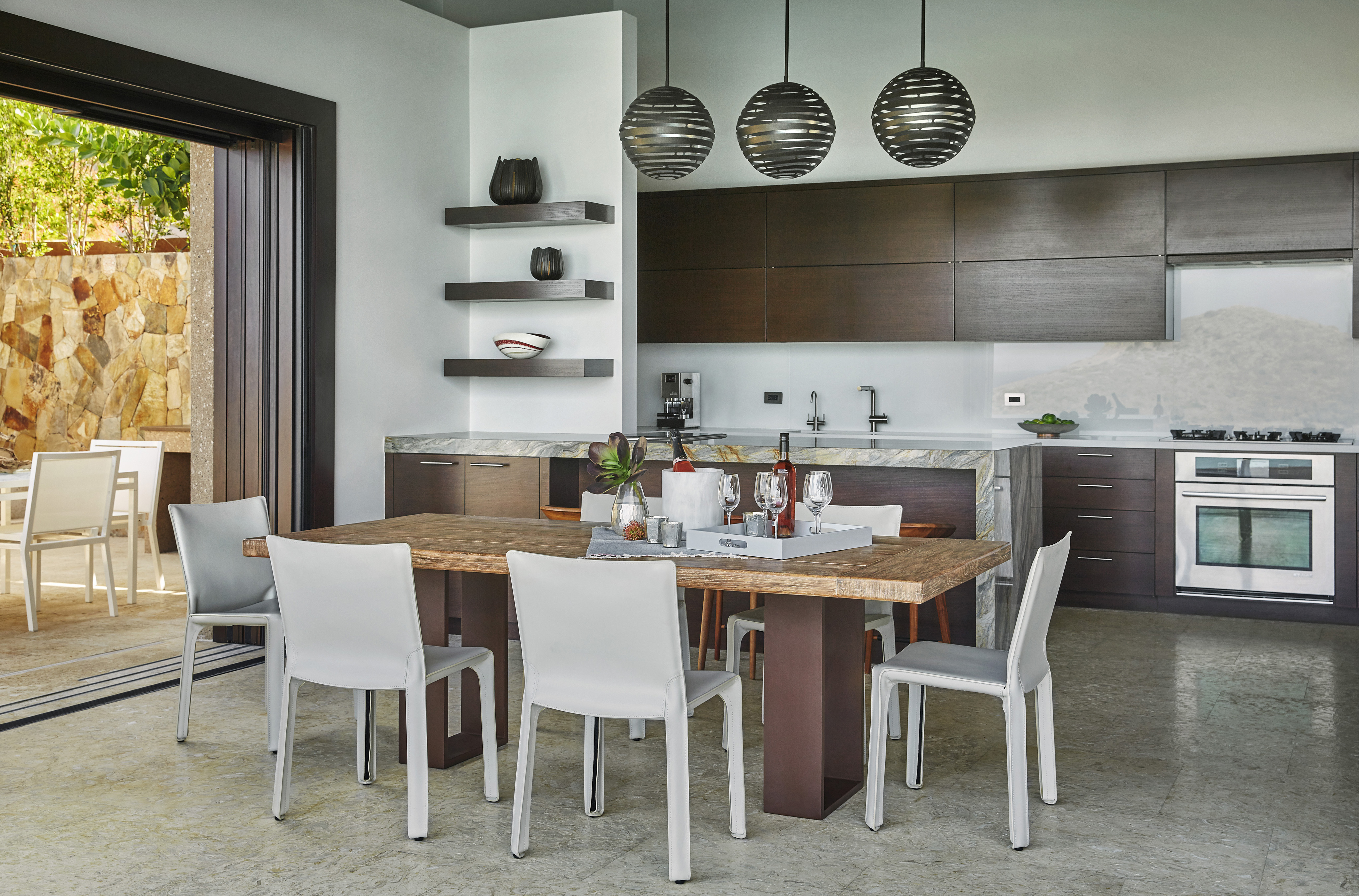 Modern kitchen and dining area in a villa with a wooden table and white chairs. The kitchen features dark brown cabinets, stainless steel appliances, and marble countertops. Large glass doors open to an outdoor patio with additional seating by the water's edge. Pendant lights hang above the table.