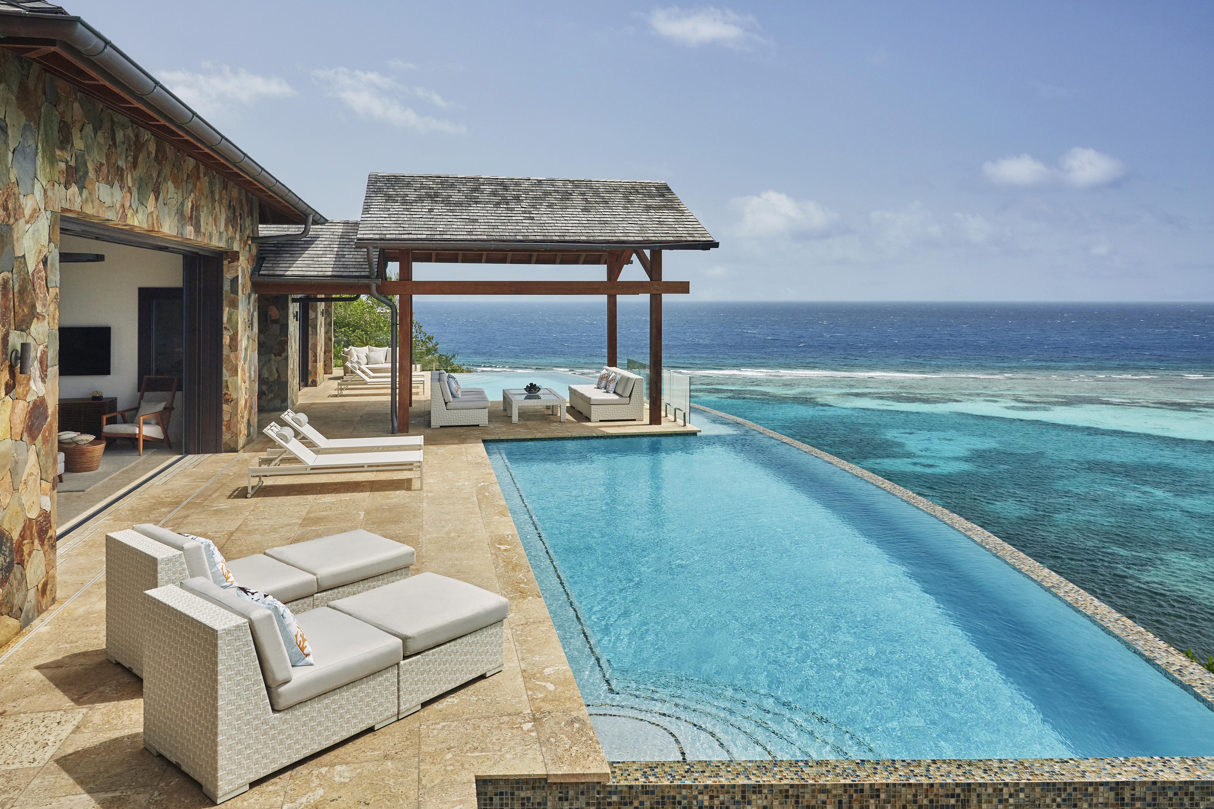 A luxurious seaside villa at the water's edge features a spacious terrace with lounge chairs and an infinity pool merging with the view of the blue ocean and sky. The villa showcases a stone facade and shaded seating area, creating a serene and opulent setting by the water.