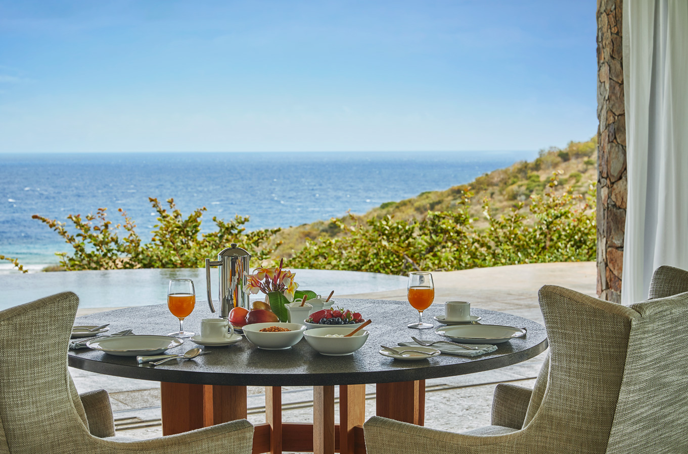 Experience in-villa dining at its finest with a round patio table set for breakfast, offering various dishes including fruit and juice. Overlooking a scenic landscape of the ocean and green hills under a clear blue sky, the cushioned chairs and tranquil atmosphere set the perfect vacation vibe.