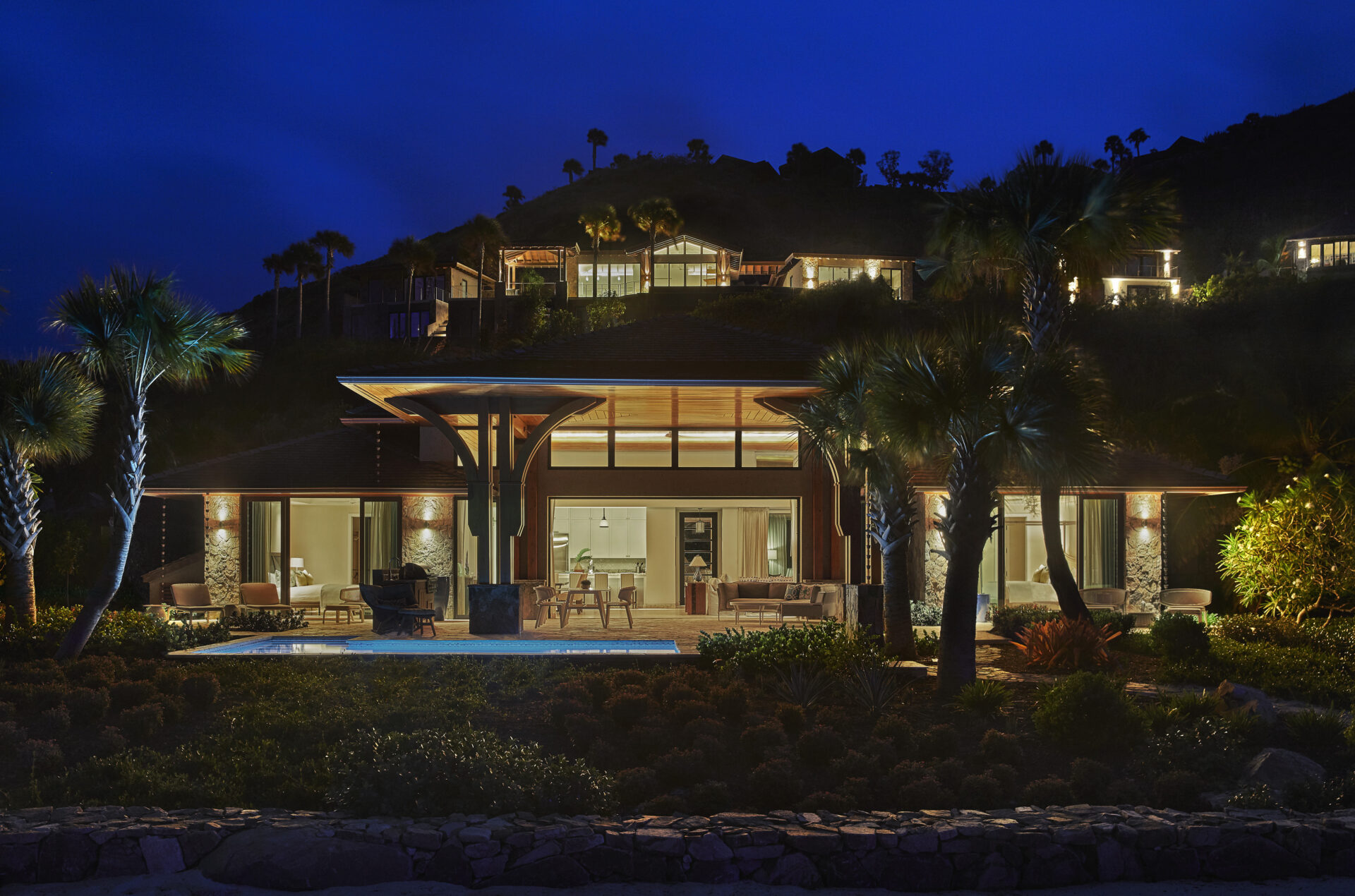 A modern, well-lit villa at night features large windows and a spacious patio. Situated on a lush hillside, surrounded by palm trees and well-maintained landscaping, it offers serene views. Other illuminated houses are visible higher on the hill.