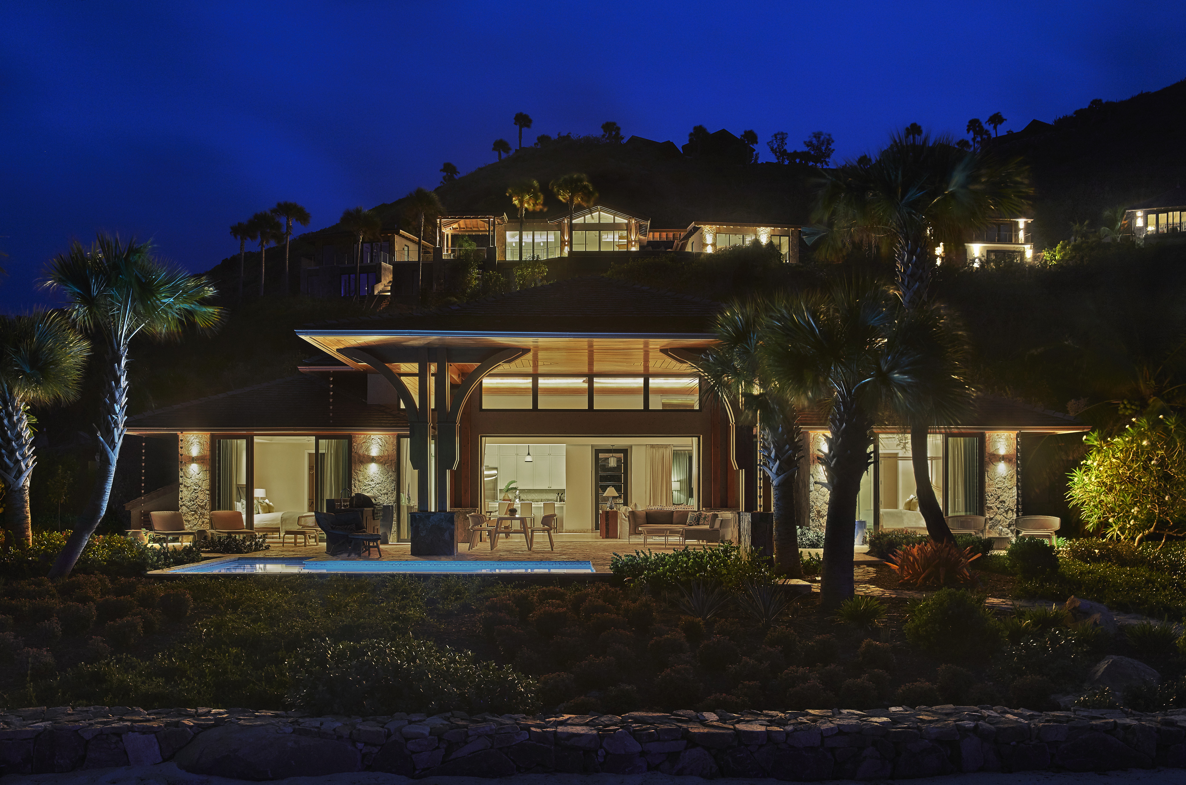 A modern, well-lit villa at night features large windows and a spacious patio. Situated on a lush hillside, surrounded by palm trees and well-maintained landscaping, it offers serene views. Other illuminated houses are visible higher on the hill.