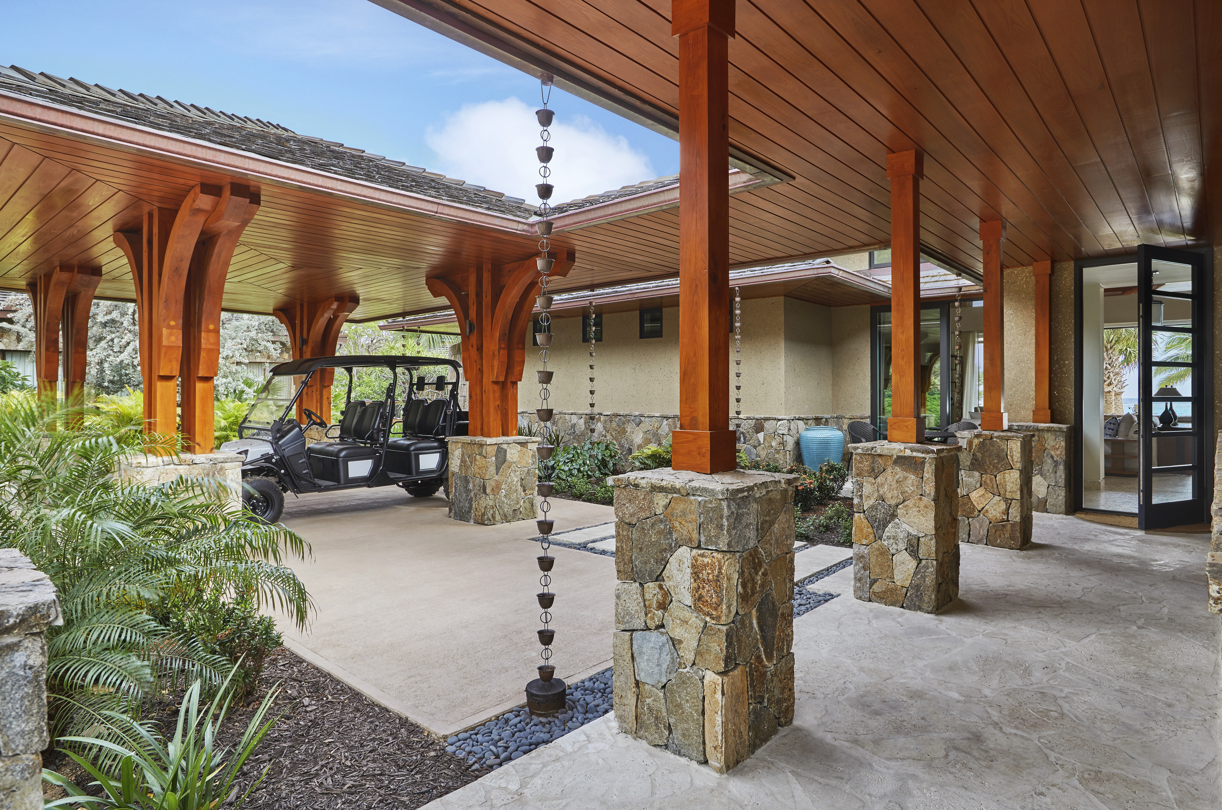 Cheemaun Villa boasts a luxurious open carport with wooden beams and stone pillars. A black golf cart rests under the elegant roof. There are hanging rain chains, tropical plants, and a clear view inside the modern house through large windows. The sky is blue with scattered clouds.