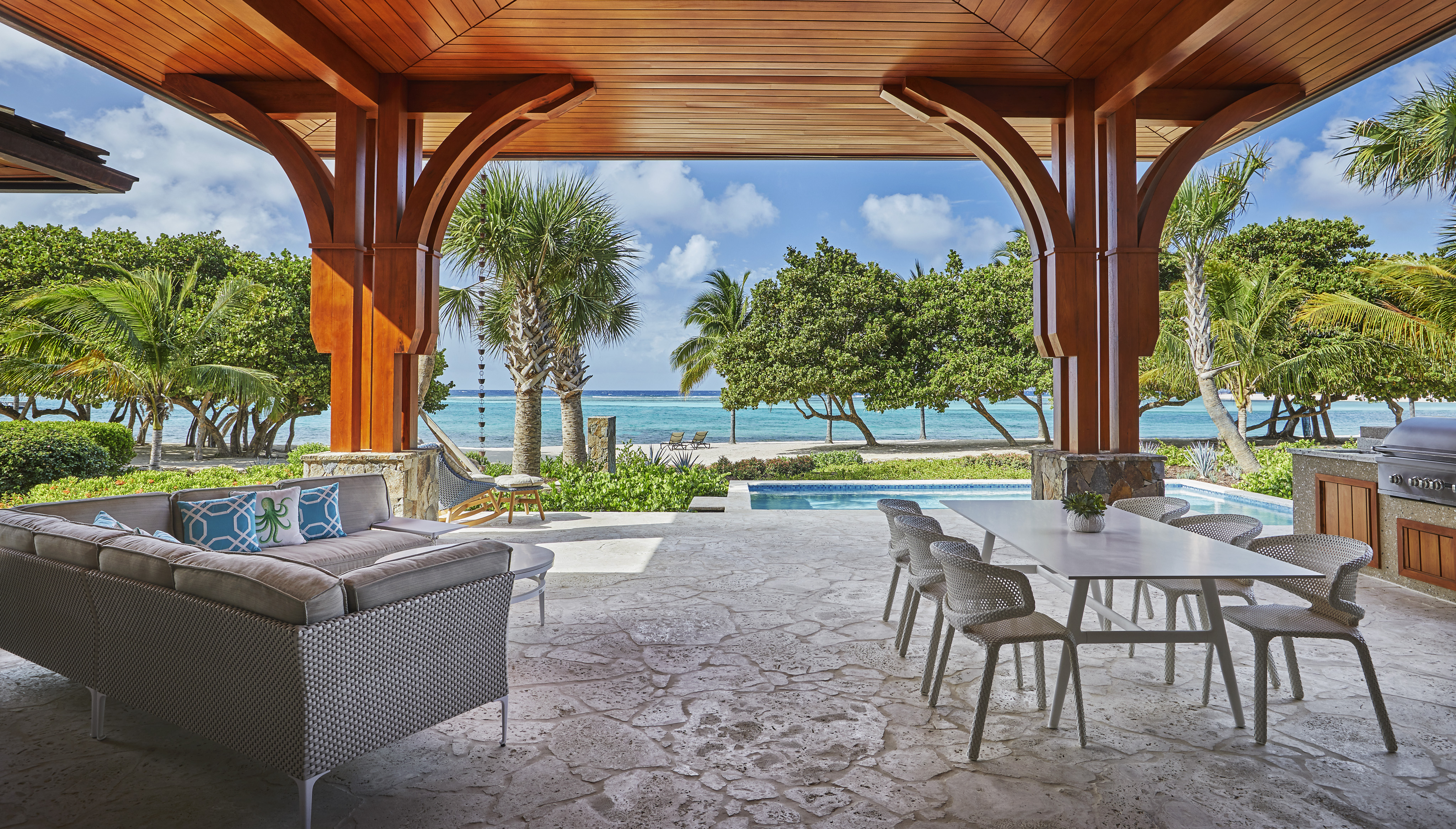 Covered outdoor patio with wooden beams and stone flooring, featuring a comfortable lounge area and dining table. The villa's patio overlooks a lush garden with palm trees, a pool, and a scenic beach view with clear blue skies in the background, like something straight out of Cheemaun.