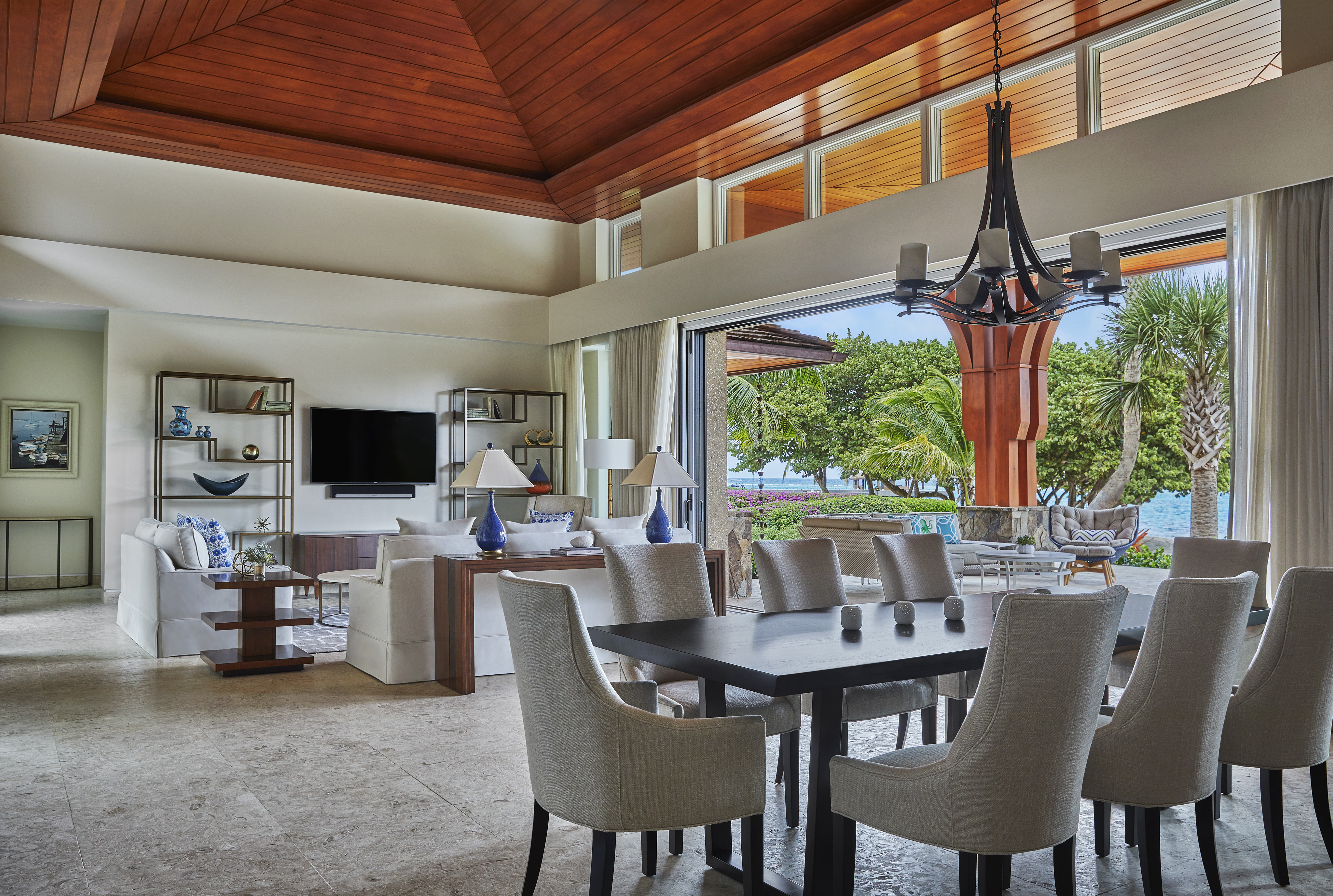 A modern, spacious living and dining area with high wooden ceilings, large windows, and sliding doors opening to a tropical outdoor view complements this exquisite villa. Furnished with white sofas, a dark wooden dining table with white chairs, and a large black chandelier, it overlooks Cheemaun gardens and a tranquil body of water.