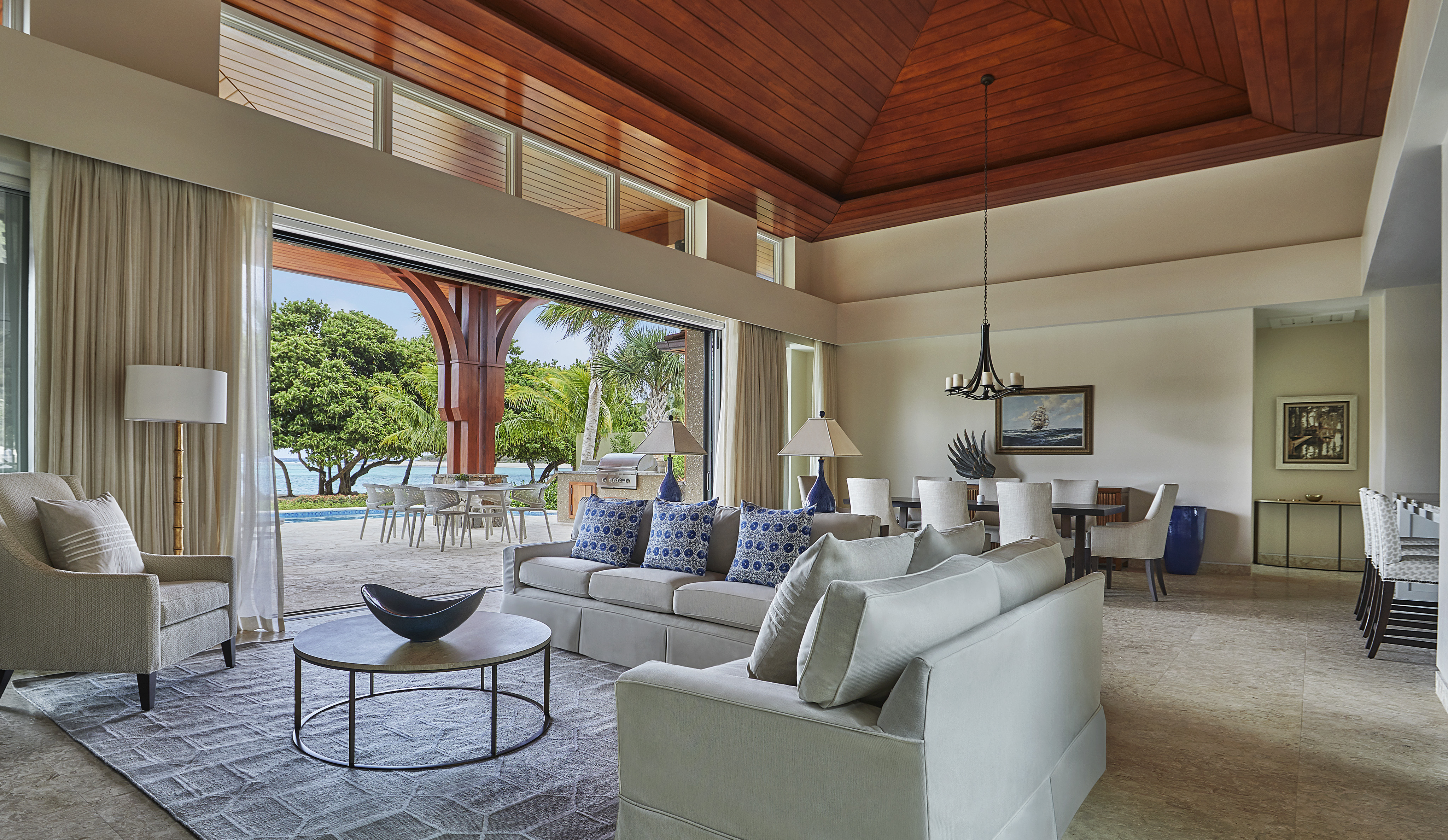 A spacious, modern living room at Cheemaun Villa features high wooden ceilings, large windows, and glass doors opening to an outdoor patio. The room boasts light-colored sofas with blue and white cushions, a central coffee table, and a dining area with a view of lush greenery outside.