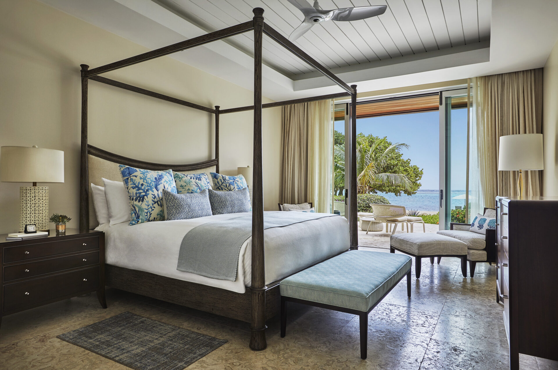 Luxurious bedroom in the Cheemaun Villa with an elegant four-poster bed adorned with blue and white pillows, matching bench at the foot, side tables with lamps, cozy seating area, and large windows showcasing a scenic ocean view surrounded by greenery.