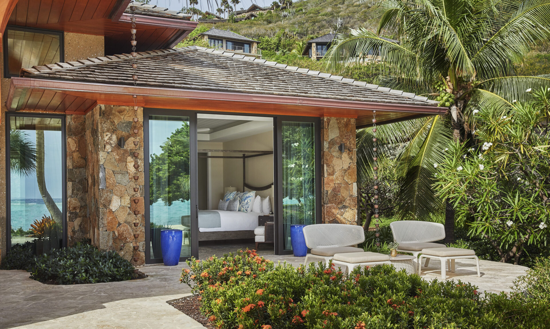 The luxurious Cheemaun Villa features a stone and wood exterior with glass sliding doors leading to a bedroom with a canopy bed. The patio outside has two lounge chairs, small tables, and is surrounded by lush greenery and palm trees with a view of the ocean.