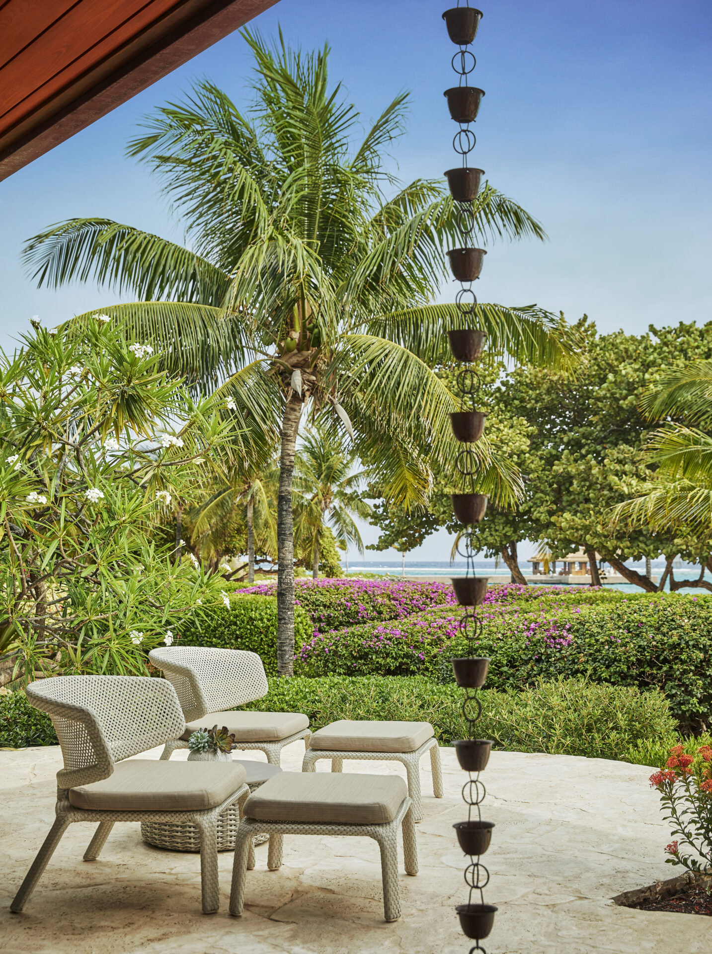 Experience Cheemaun Villa's tropical outdoor seating area featuring two wicker chairs, matching ottomans, and a side table with a potted plant. Enjoy the lush greenery, palm trees, and ocean views in the distance. A vertical rain chain adds to the serene ambiance.