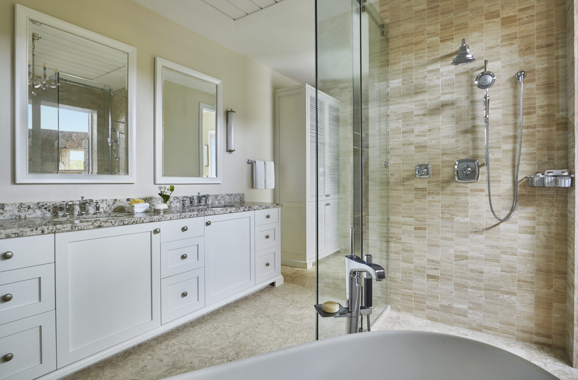 Modern bathroom in Cheemaun Villa with a dual-sink white vanity, granite countertop, and large glass-enclosed shower with beige tiles. Two mirrors hang above the sinks, and a freestanding bathtub sits in the foreground. A towel rack and wall-mounted light fixture complete the look.