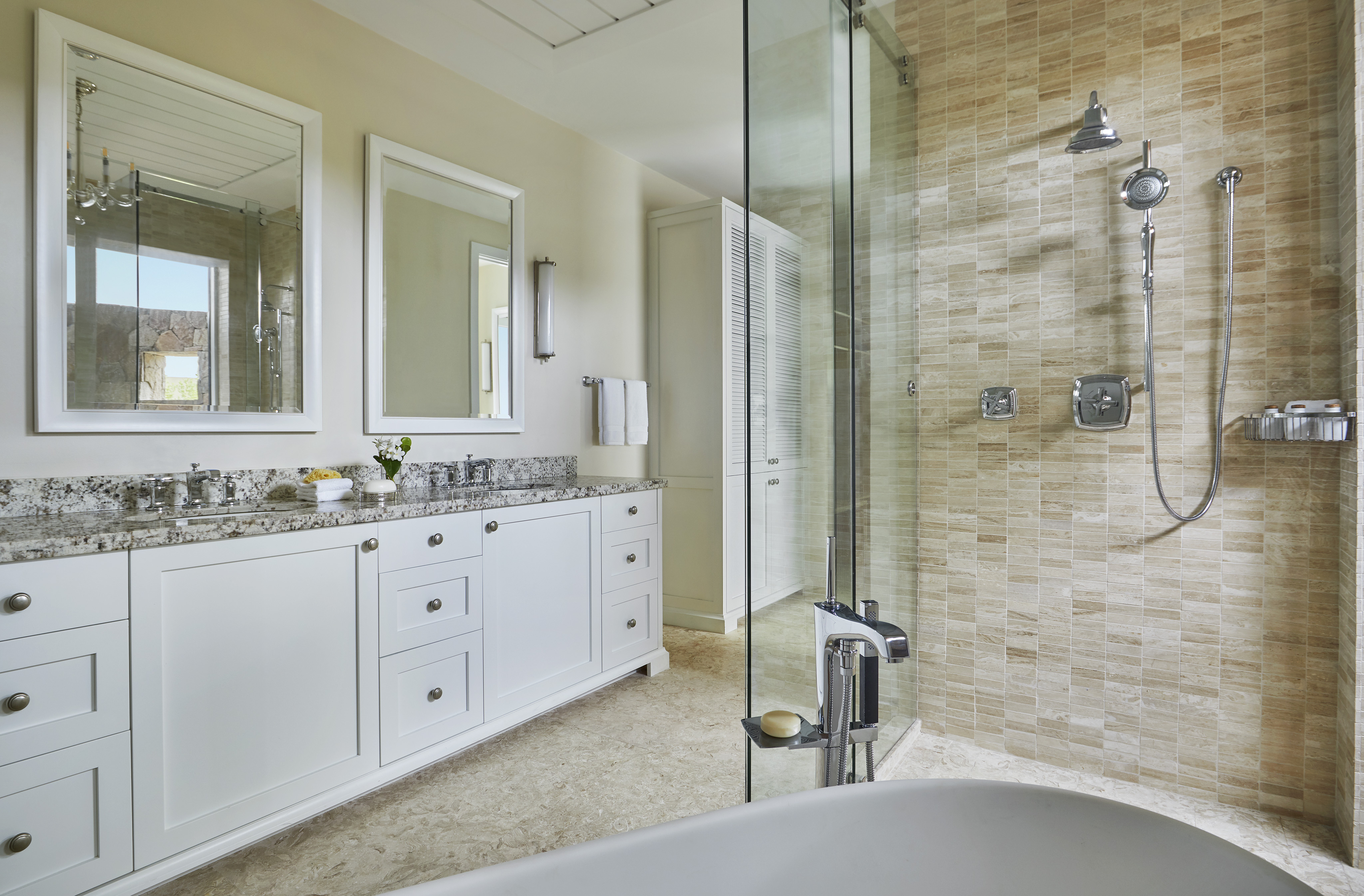 Modern bathroom in Cheemaun Villa with a dual-sink white vanity, granite countertop, and large glass-enclosed shower with beige tiles. Two mirrors hang above the sinks, and a freestanding bathtub sits in the foreground. A towel rack and wall-mounted light fixture complete the look.