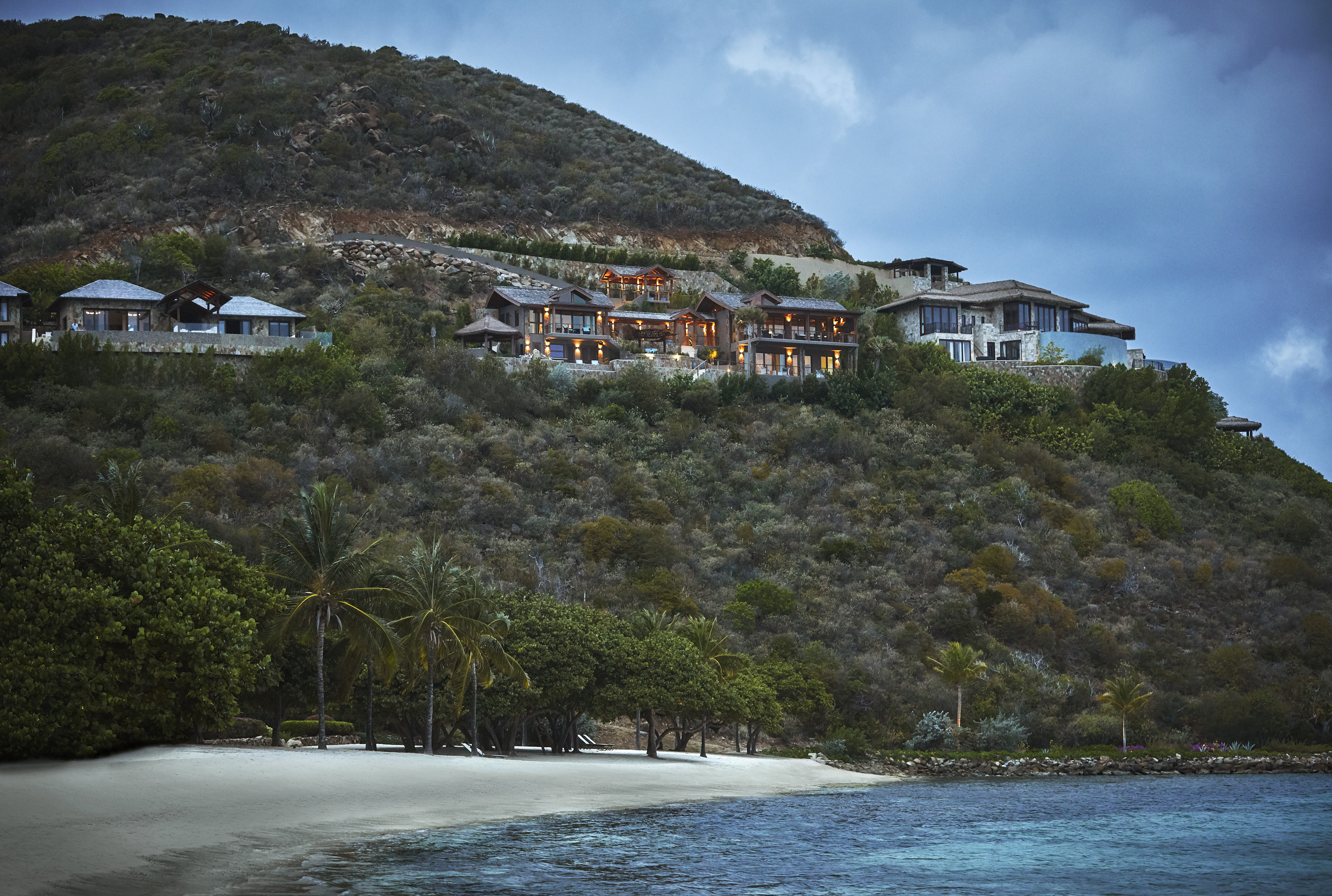 Poseidon's Perch Villa offers luxury hillside living with breathtaking views of a sandy beach and clear blue waters. The homes are nestled amid lush green foliage, with palm trees lining the shore. The scene is tranquil, with a cloudy sky adding a serene atmosphere to the picturesque landscape.