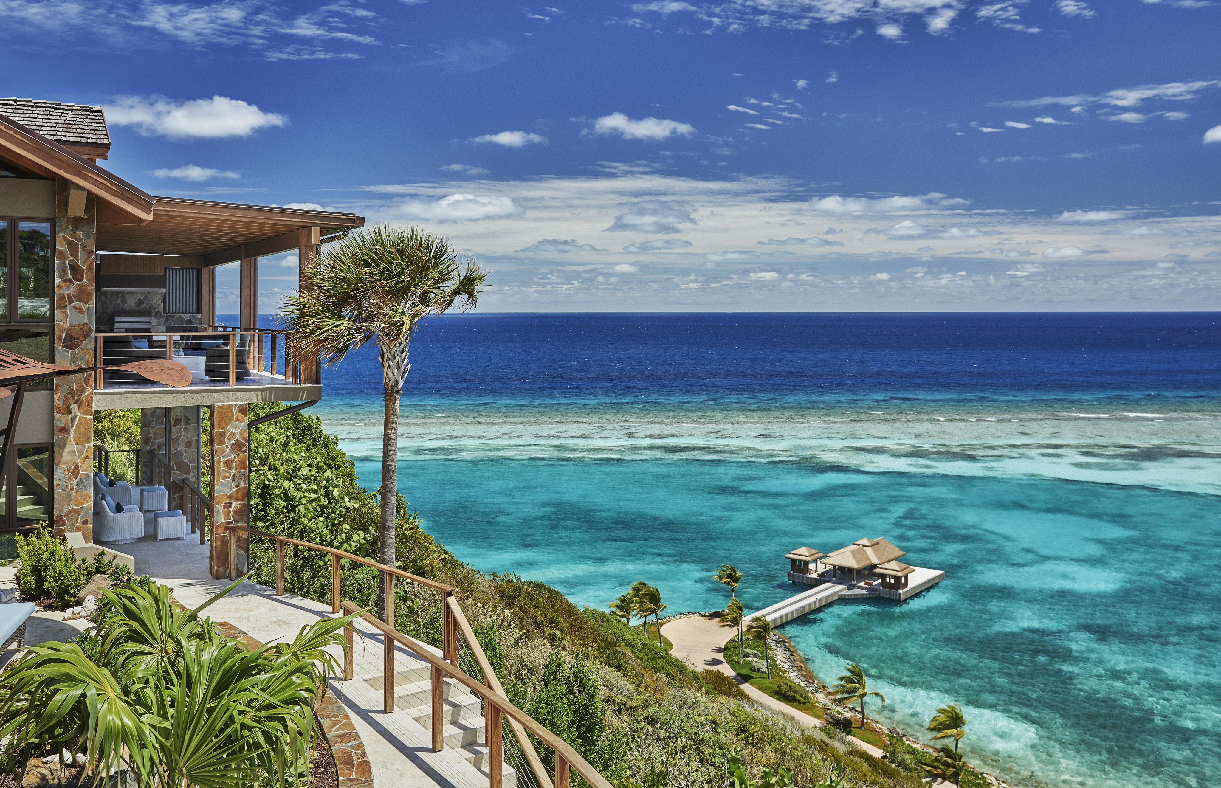 Poseidon's Perch Villa is a luxurious oceanside retreat with a spacious balcony overlooking pristine blue sea. A small wooden pavilion stands at the end of a white pier, surrounded by lush greenery and palm trees. The crystal-clear water features varying shades of blue and turquoise.