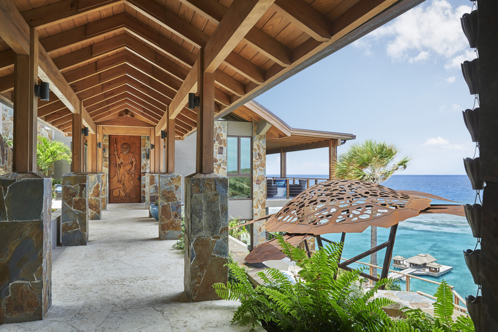 A scenic outdoor walkway with a wooden roof and decorative beams overlooks the ocean at Poseidon's Perch Villa. Stone columns support the structure, and a carved wooden door is visible at the end of the path. Lush green ferns and a sculptural element add to the tropical ambiance of this luxury beachfront property.