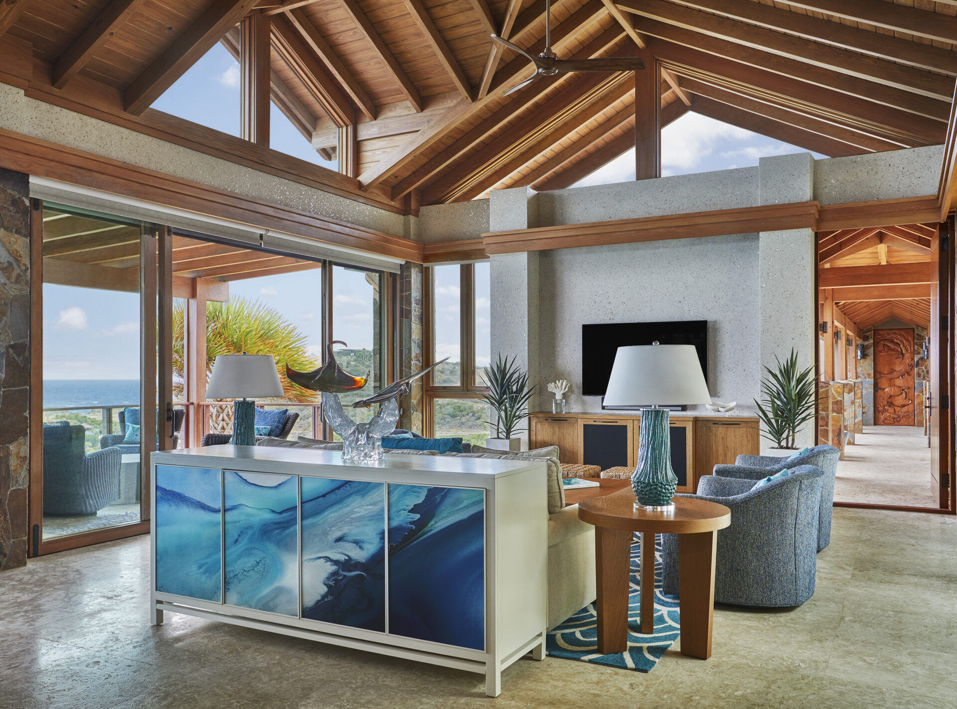 Poseidon's Perch Villa offers a luxurious living space with wooden vaulted ceilings and large windows revealing an ocean view. The room features blue upholstered furniture, a TV, and a white sideboard adorned with blue abstract art. A table lamp and decorative objects are placed on the sideboard and tables.