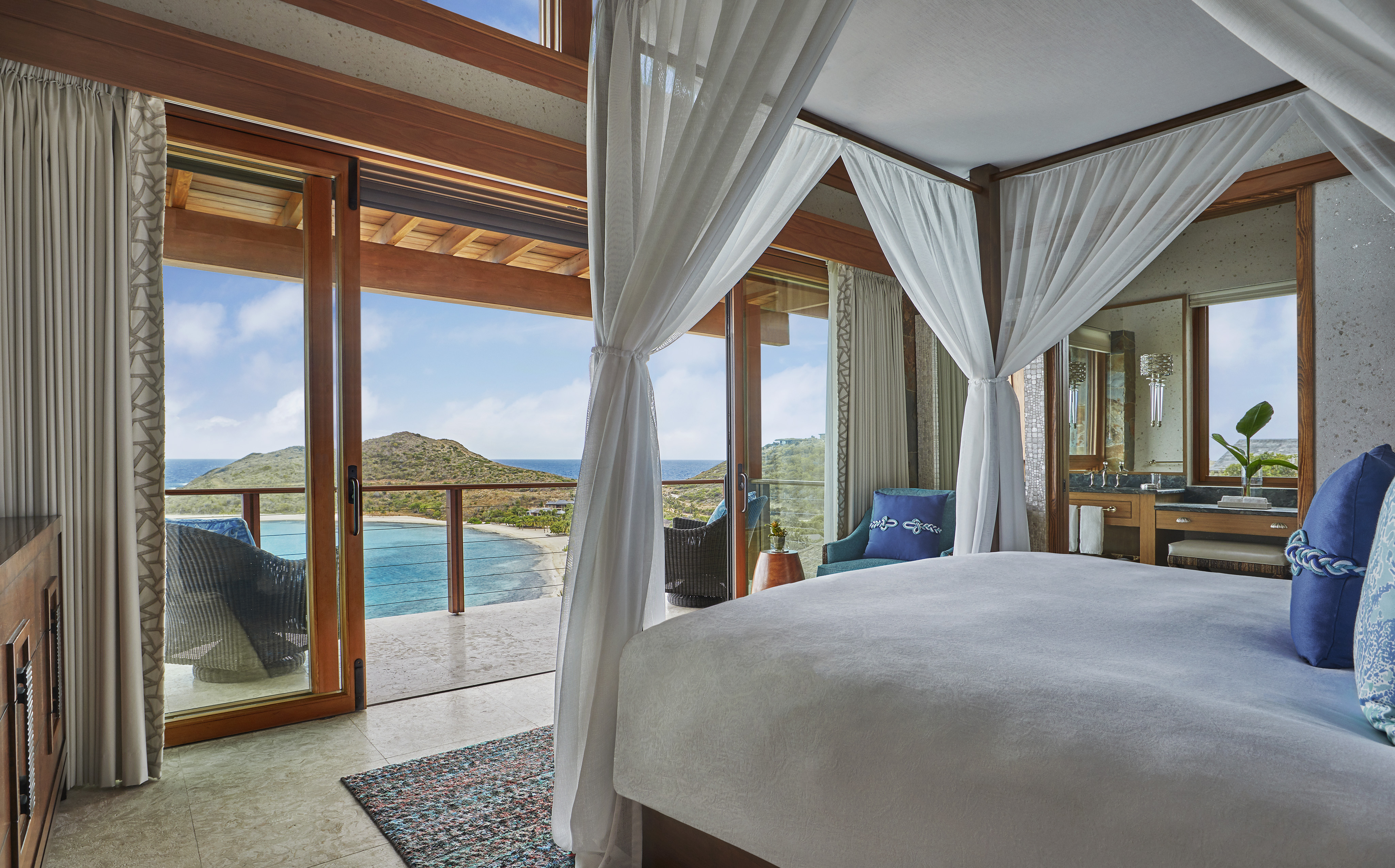 A luxurious bedroom at Poseidon's Perch Villa boasts a canopy bed draped in white curtains, overlooking a scenic ocean view through large glass doors. Elegantly decorated with blue accents, the room includes a cozy balcony with seating. In the distance, a lush island is visible.