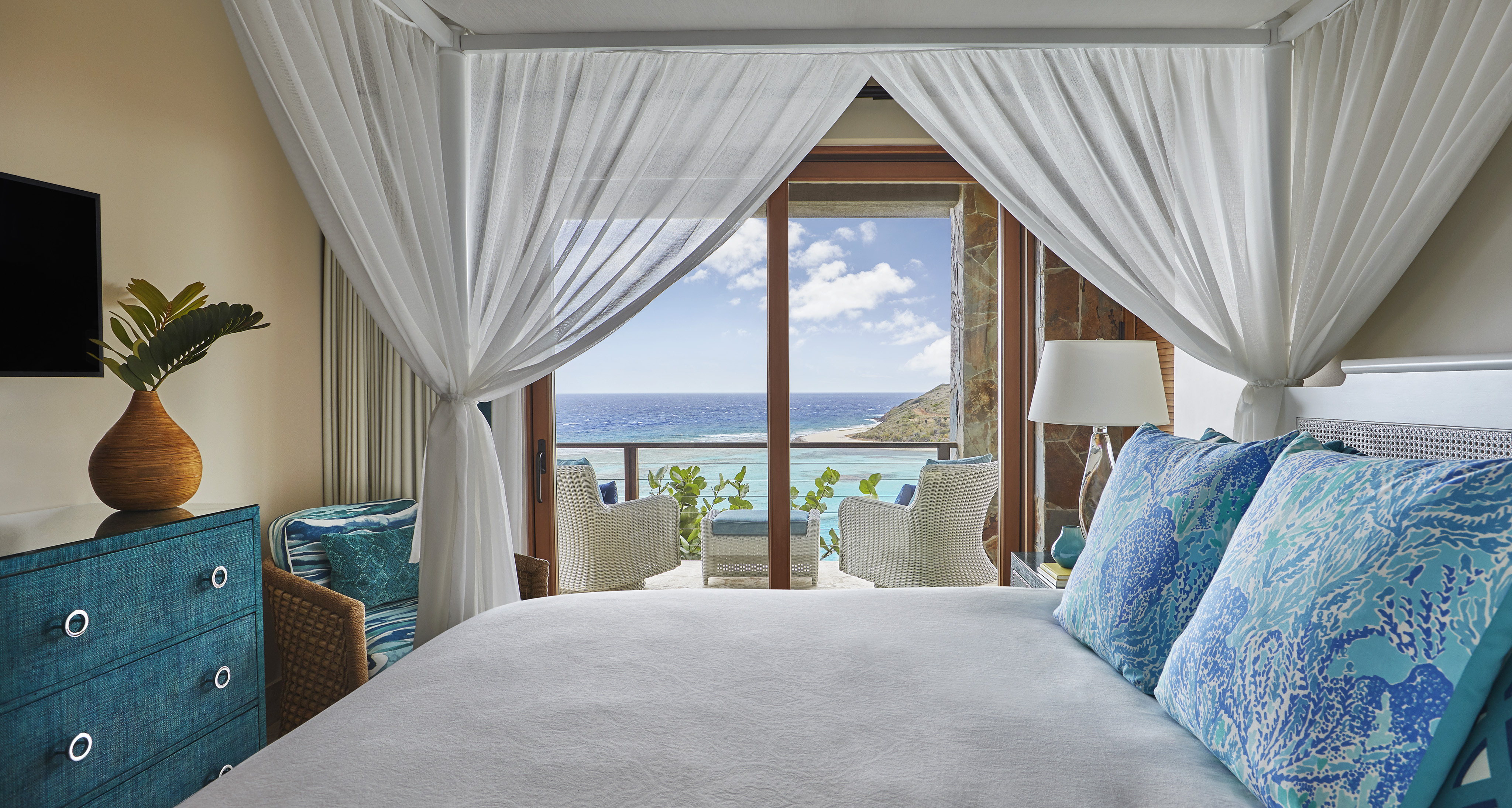 A luxurious bedroom in Poseidon's Perch villa, featuring a four-poster bed covered in white linens and decorative blue pillows. The room boasts a turquoise dresser, a flat-screen TV, and wicker chairs. Sliding doors open to a balcony with white chairs, offering a panoramic ocean view.