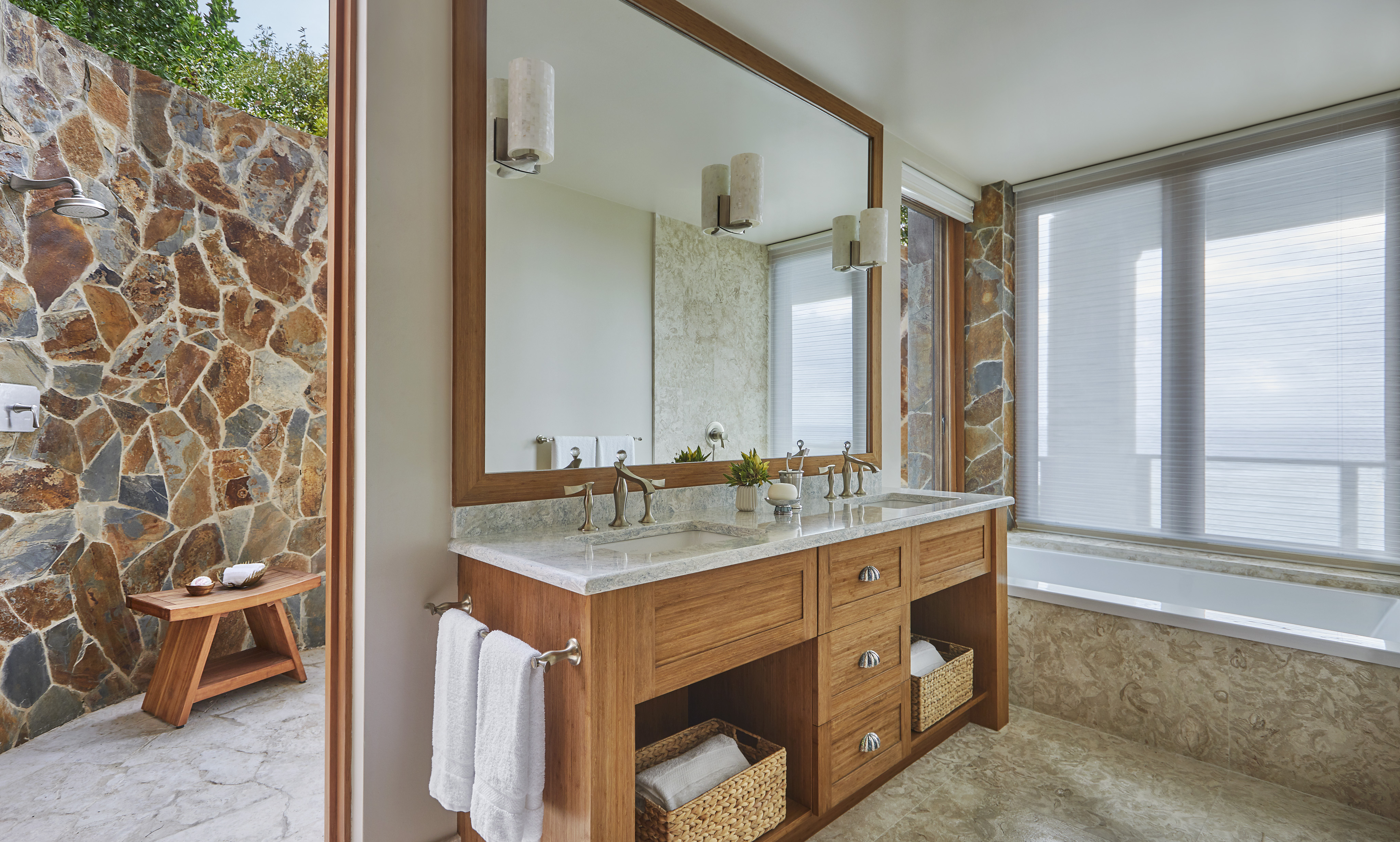 In Poseidon's Perch Villa, a modern bathroom features a large wooden vanity with a marble countertop, dual sinks, and a wide mirror. Below the sinks, two wicker baskets rest on a lower shelf. The room boasts a glass-enclosed shower with stone walls and a window next to the bathtub.