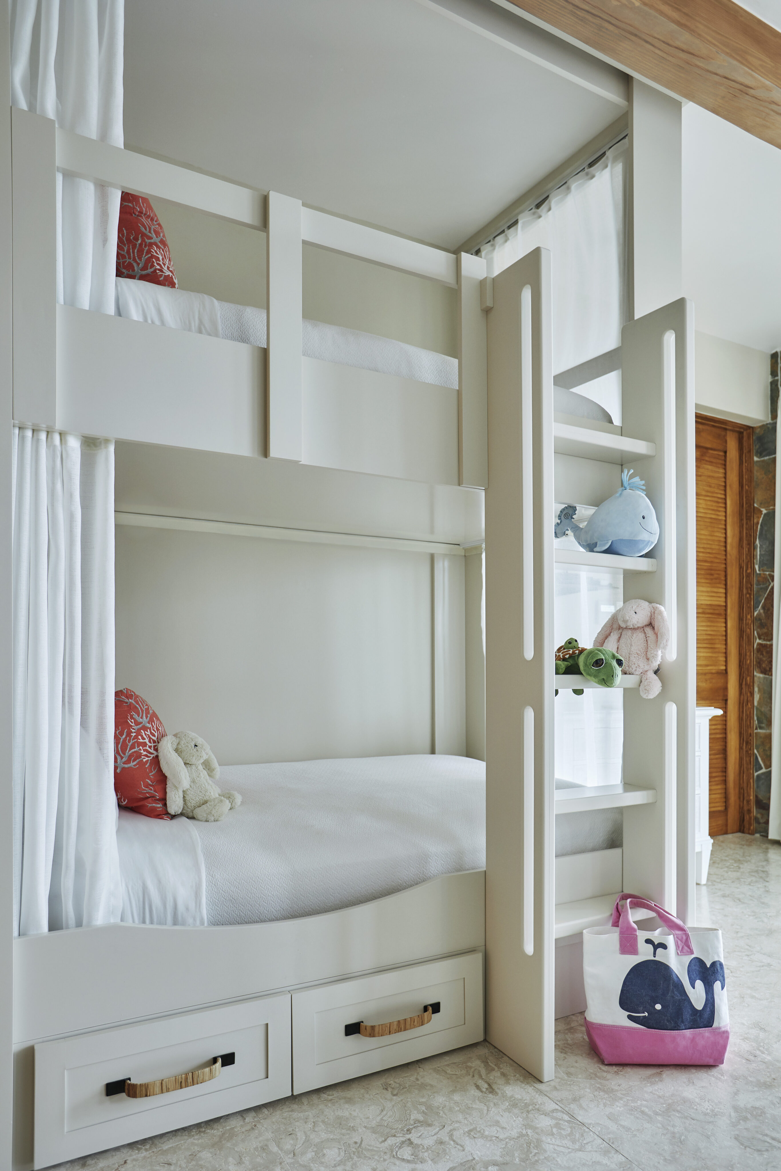 A cozy children's bedroom, aptly named Poseidon's Perch, features white bunk beds with built-in storage drawers underneath. The top bunk is accessed by a ladder. Plush toys, a pink whale bag, and red pillows with white patterns add playful touches to the bright and airy villa-like space.