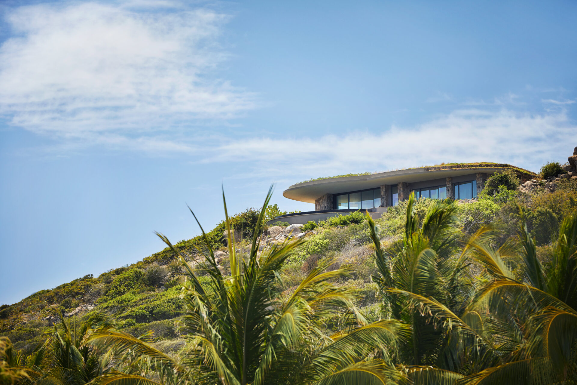 Rainbow Villa, a modern, minimalist house with large glass windows, is nestled into a lush, green hillside under a partly cloudy sky. Palm trees in the foreground frame the scene beautifully. The villa appears to blend seamlessly with the natural surroundings.