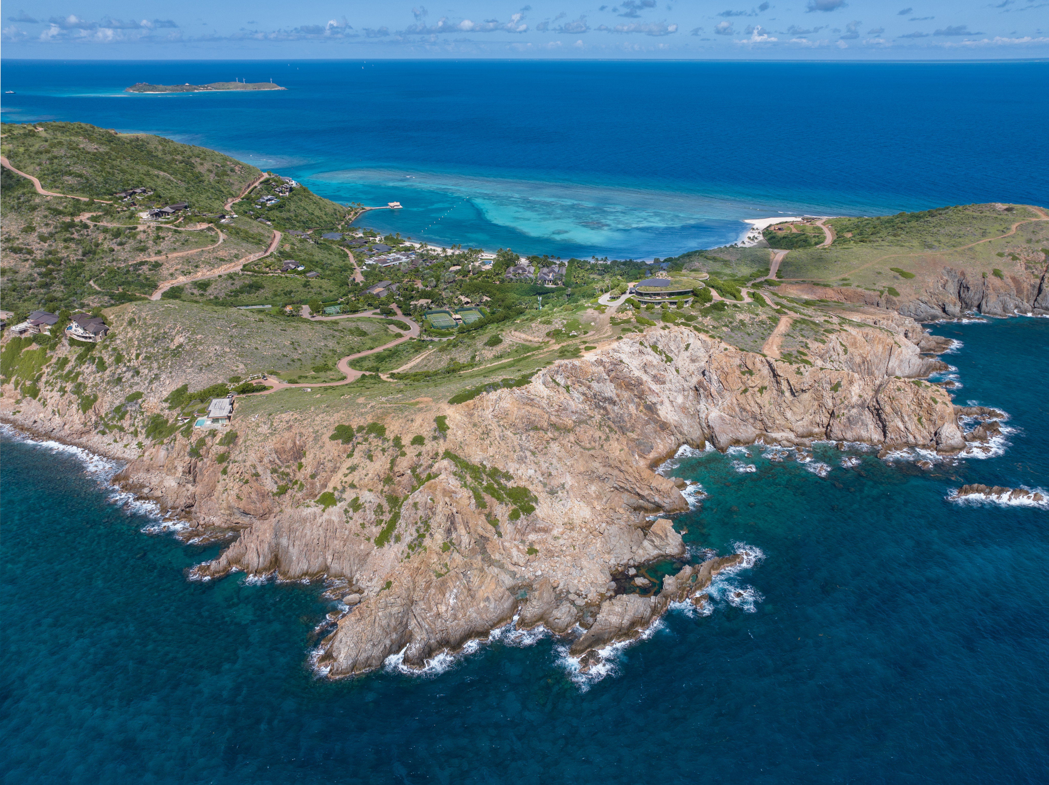 Aerial view of a rugged coastline with rocky cliffs and green hills above a deep blue ocean. Serpentine roads wind through scattered houses on the hilly landscape, part of the majestic Atlantic Ridge. Turquoise waters and a distant island are visible in the background under a partly cloudy sky.