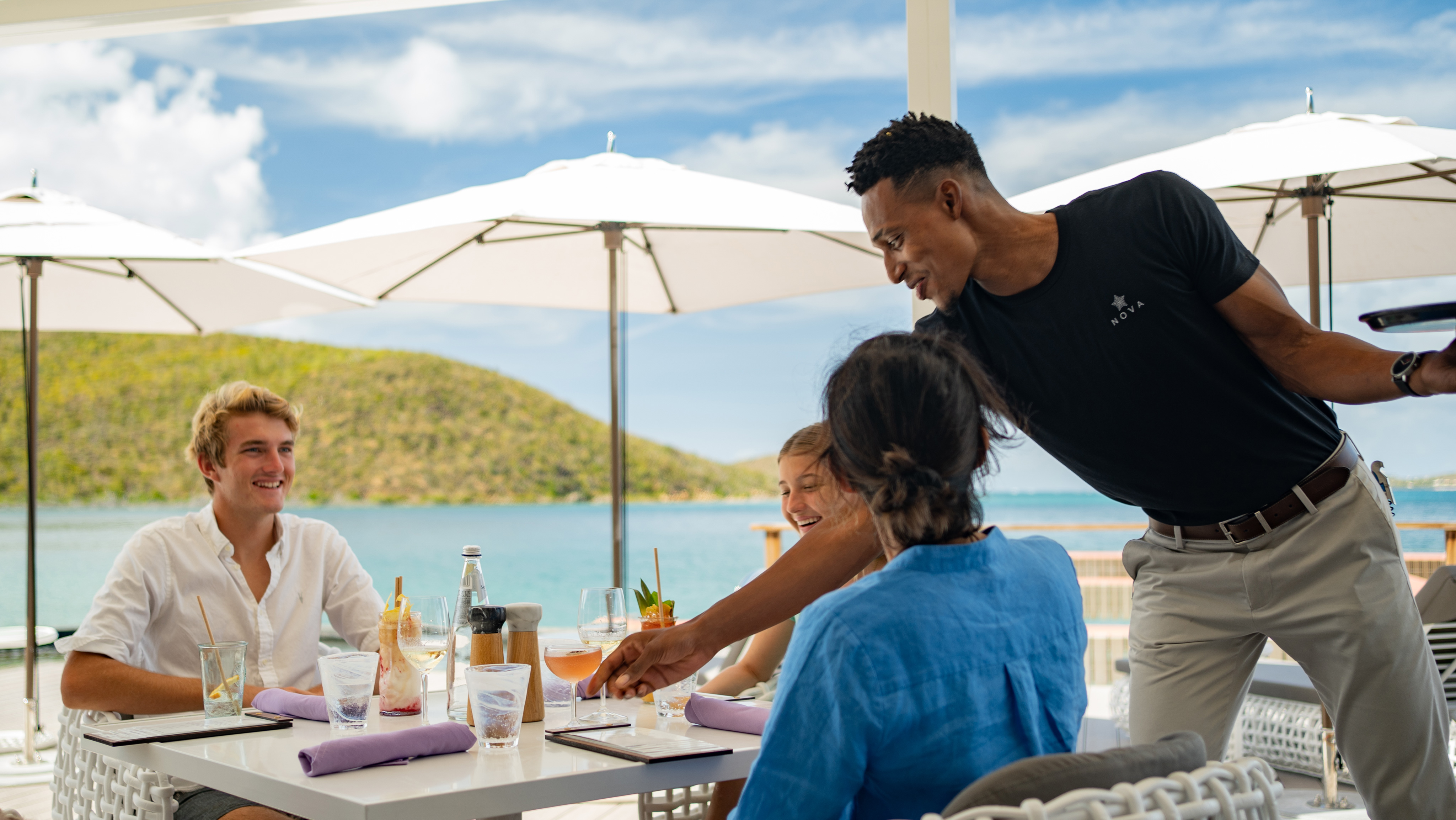 A waiter serves Nova cocktails to a group of three at an outdoor seaside restaurant. They relax under white umbrellas, with scenic hills and clear skies painting the backdrop. All appear to be savoring both the drinks and the sunny day.