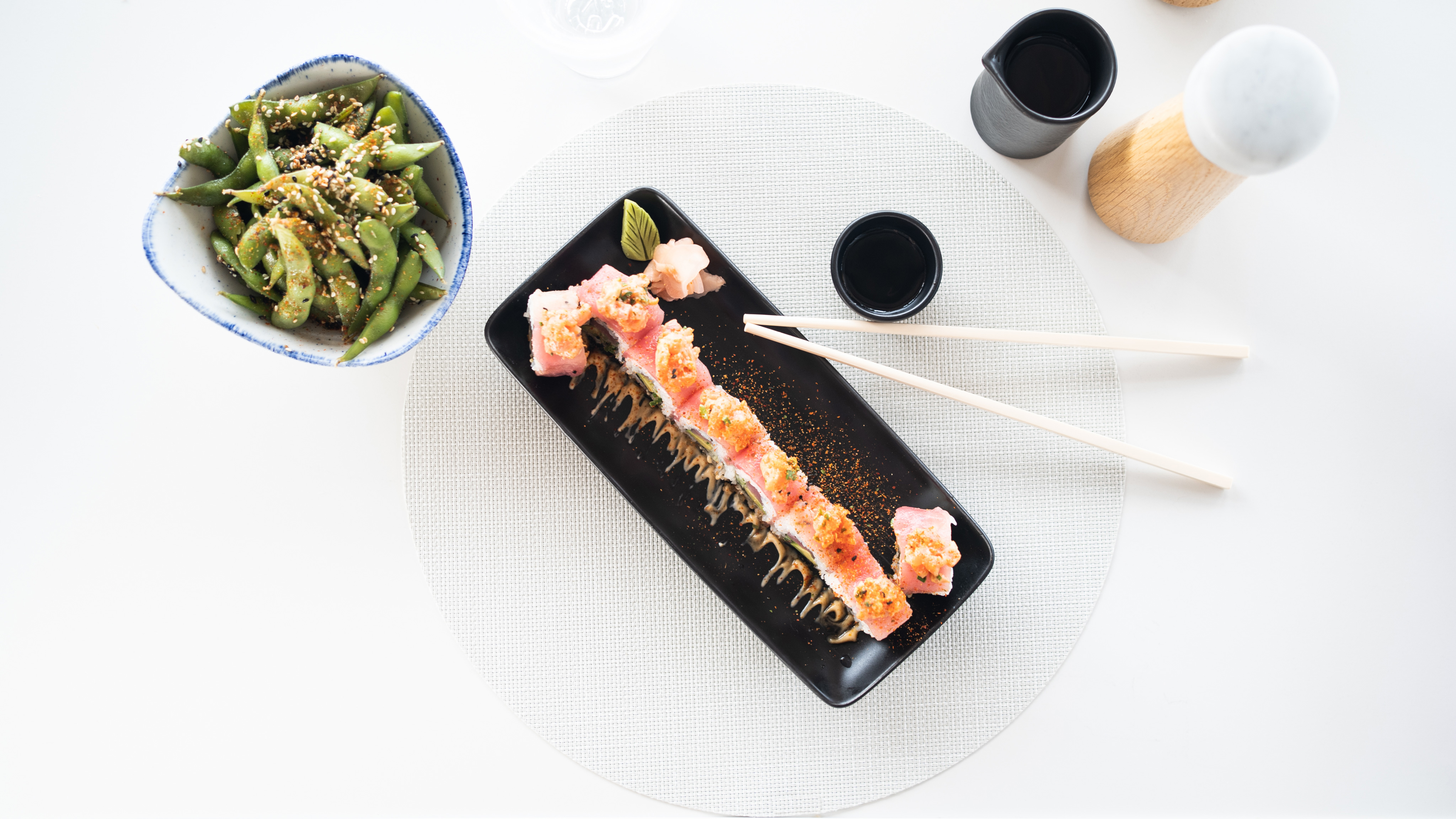 A top-view of a sushi platter on a black rectangular dish with chopsticks, alongside a bowl of seasoned edamame on a white circular placemat. A light wood pepper grinder and sauce cups are seen nearby against a Nova-white background.