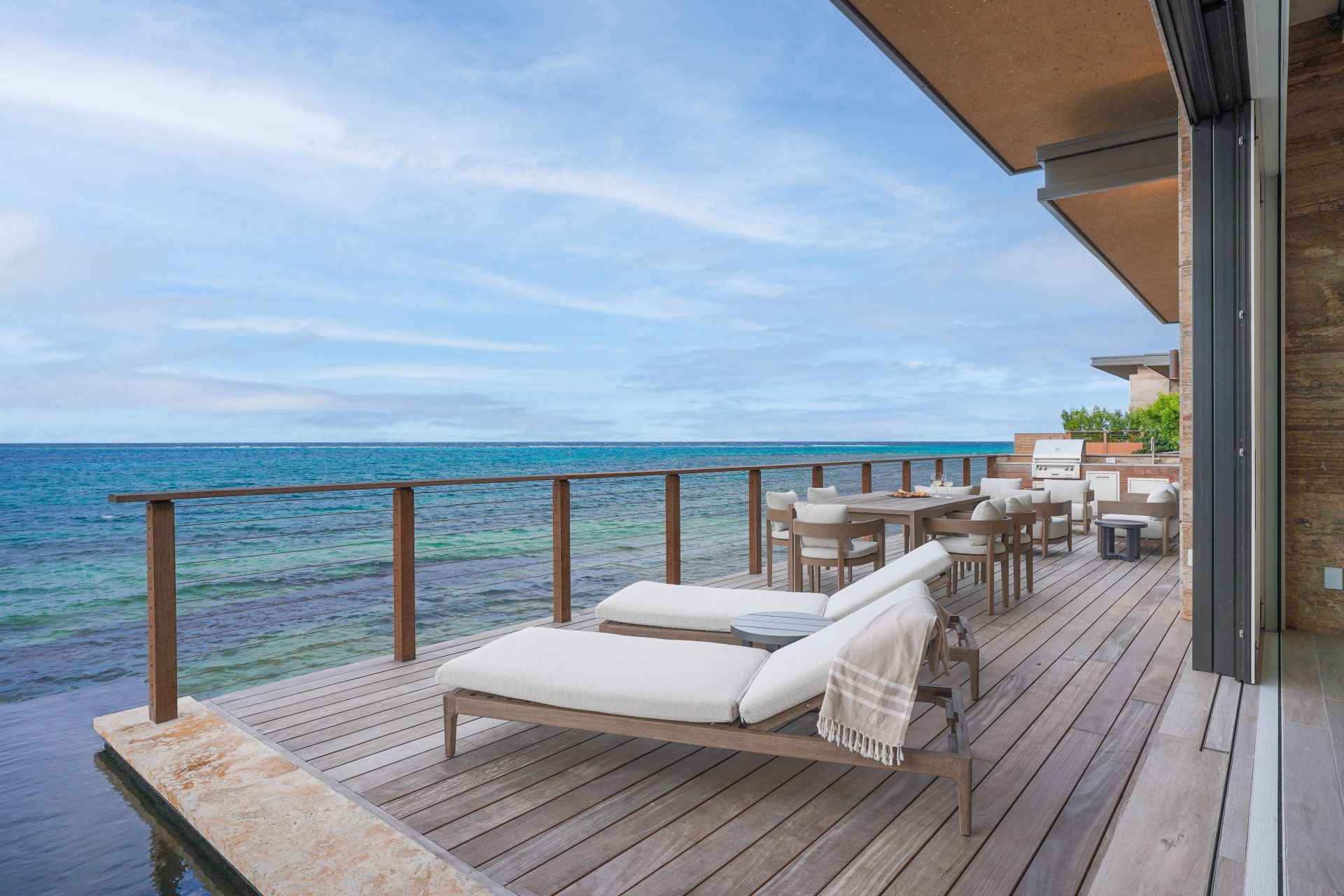 A wooden deck juts out over the ocean, offering a picturesque view at Ocean Villa OV2. Two cushioned chaise lounges with a blanket draped on one provide a relaxing seating area, while a wooden table with chairs is set up nearby. Clear blue sky and calm waters complete the tranquil scene.