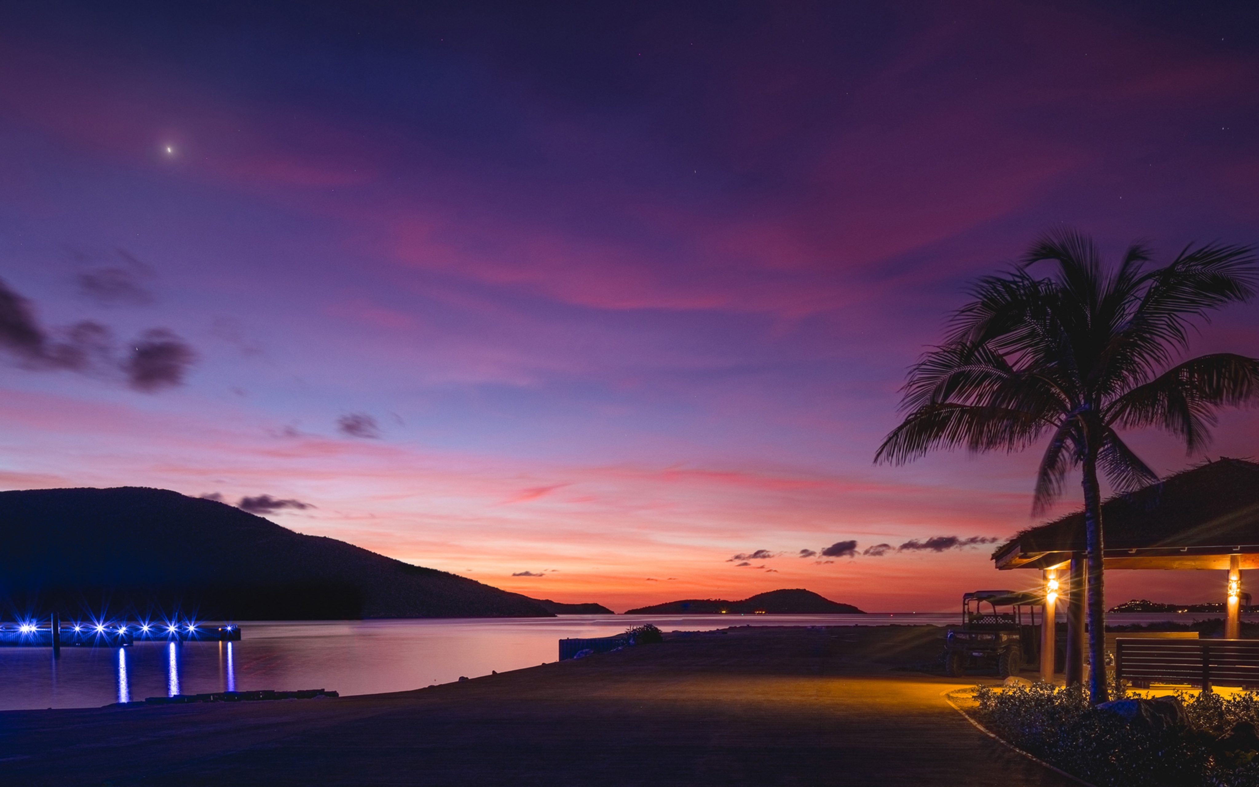 A serene beach scene at dusk with vibrant purple and orange hues in the sky. A silhouette of a palm tree and a building is on the right, while a faintly lit pier extends into calm water on the left. Hills are visible in the distance under a twinkling star, perfect for resort vacation packages in the BVI.