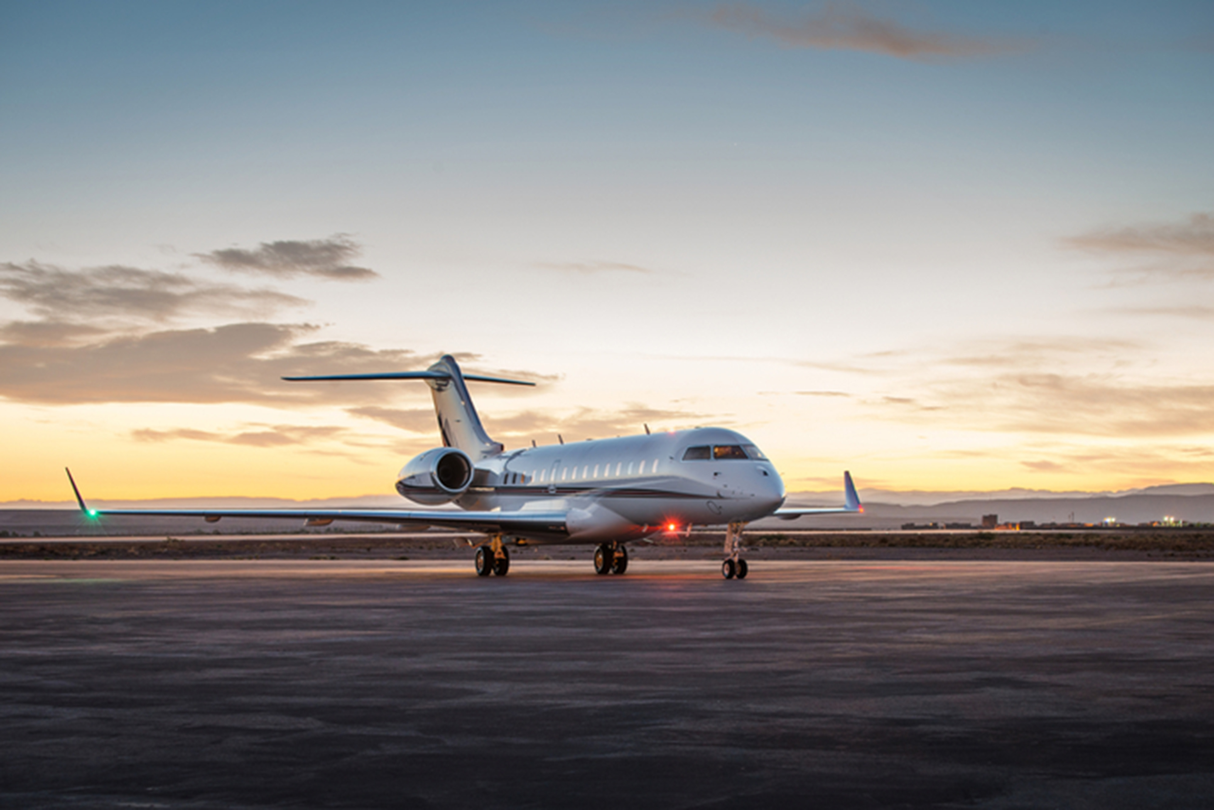 A sleek private jet from NetJets sits on an empty tarmac under a striking sunset sky. The horizon glows with warm orange and pink hues, while the aircraft's lights are on, indicating it's ready to fly. Sparse clouds and distant mountains complete the serene backdrop.