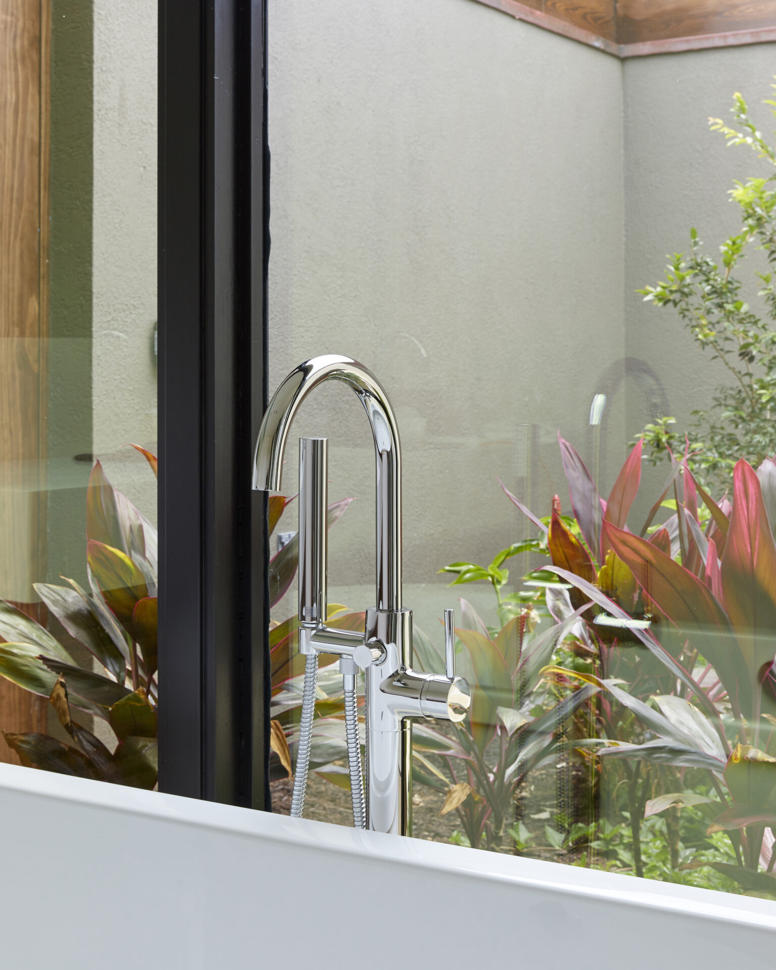 A modern, chrome bathroom faucet is mounted next to a large window with a view of an outdoor garden. The garden features a variety of green and reddish plants. The clean, sleek faucet contrasts against the natural backdrop, creating a serene and stylish ambiance reminiscent of a beach house retreat.