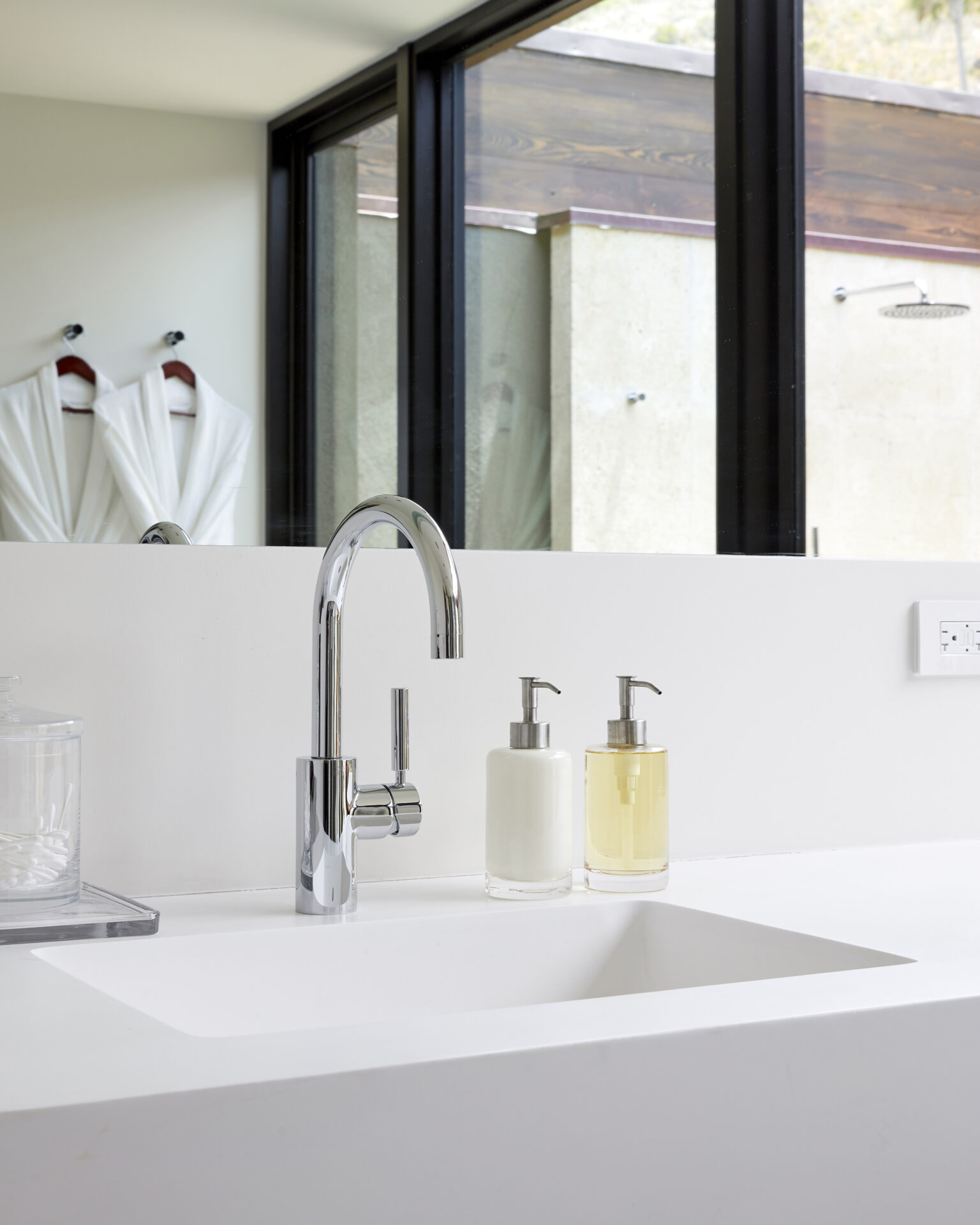 A modern bathroom in a beach house features a sleek, white countertop with a built-in sink. A chrome faucet is mounted on the countertop alongside two soap dispensers, one with white liquid and the other with yellow. In the background, a glass wall reveals two white robes hanging.