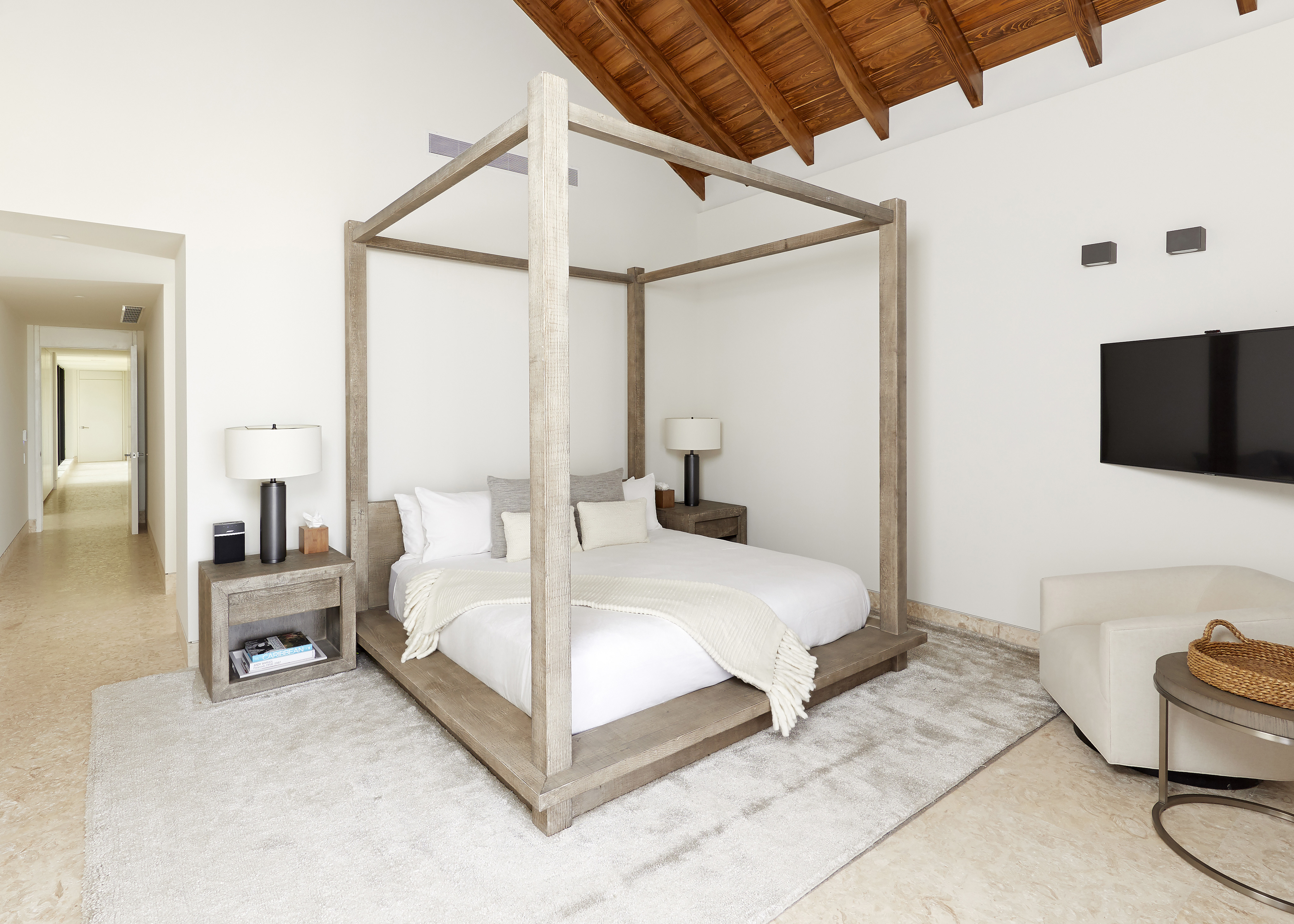 A modern bedroom in a seaside rental with a large four-poster bed, white and neutral bedding, two side tables with lamps, and a wall-mounted television. The room features a sloped wood ceiling, beige walls, and a hallway leading to other areas. A cozy armchair and basket are in the corner.