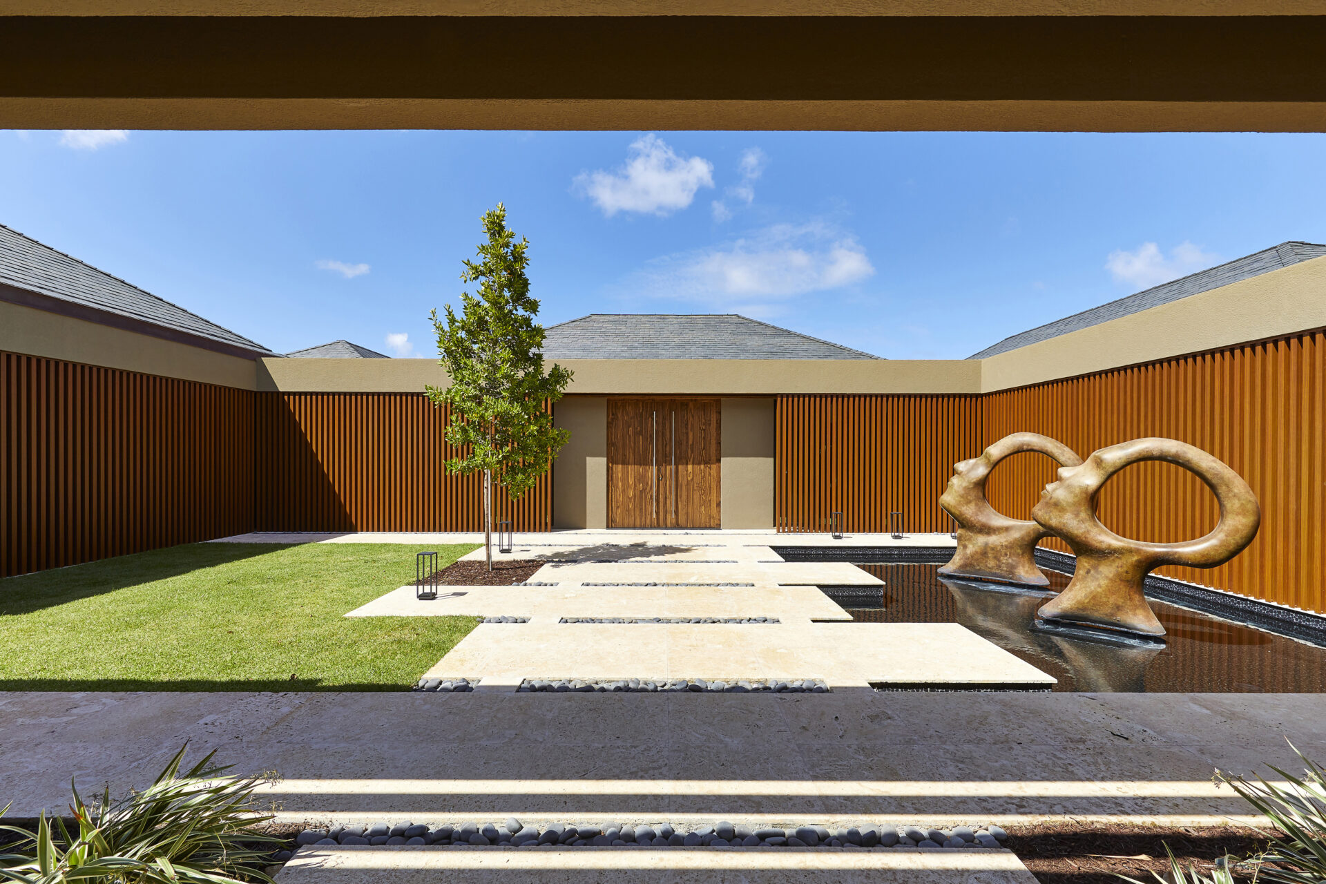 A modern courtyard at the beach house features a minimalist design with paved stepping stones over a reflecting pool, two large abstract bronze sculptures, a central tree, and contemporary wooden walls. In the background, double wooden doors and a blue sky with a few clouds are visible.