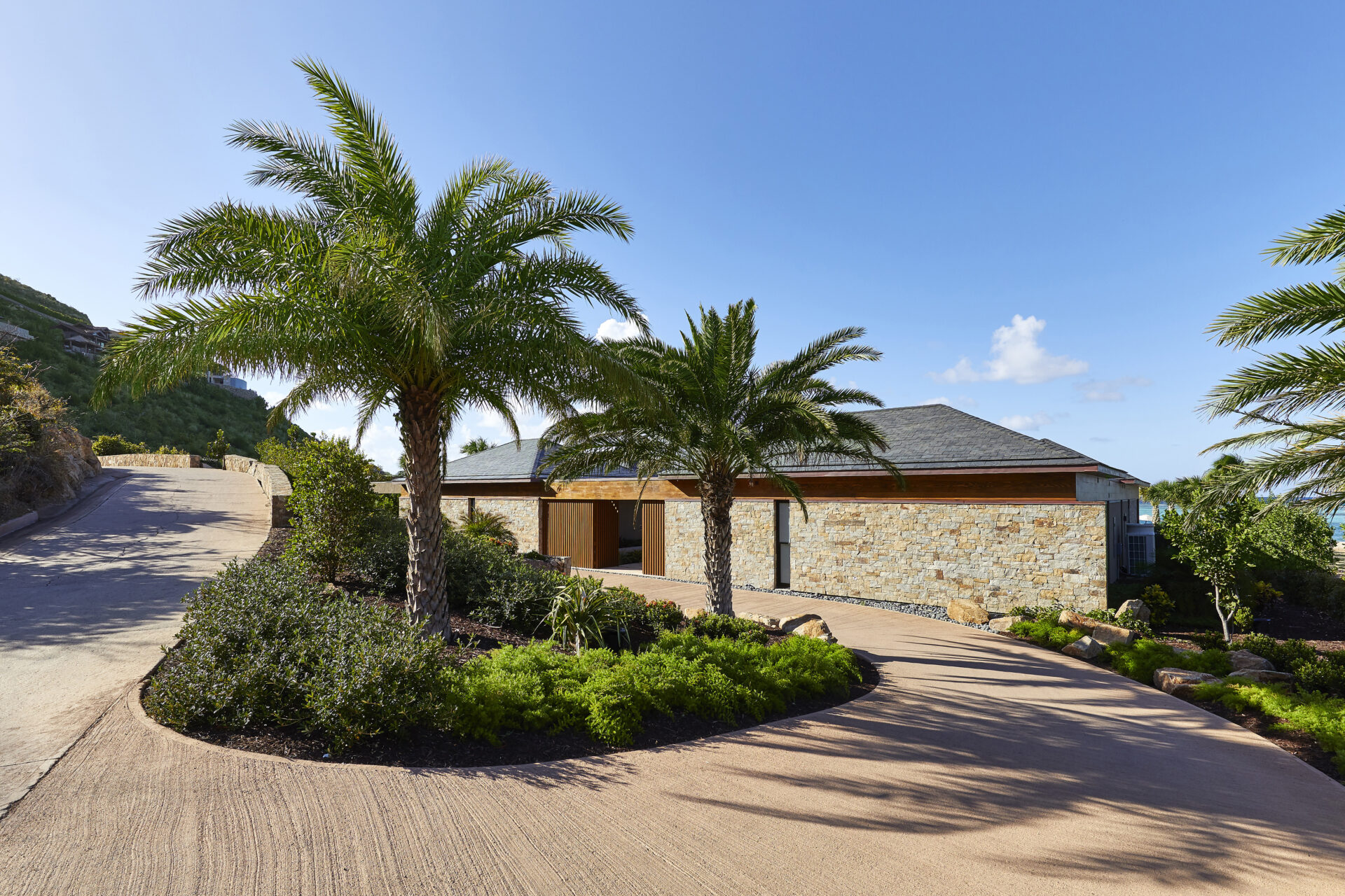 A modern, single-story stone beach house with a sloping roof is surrounded by lush greenery. Two tall palm trees grace the driveway, which winds along a gently curving path. The sky is clear and blue, creating a serene and inviting atmosphere.