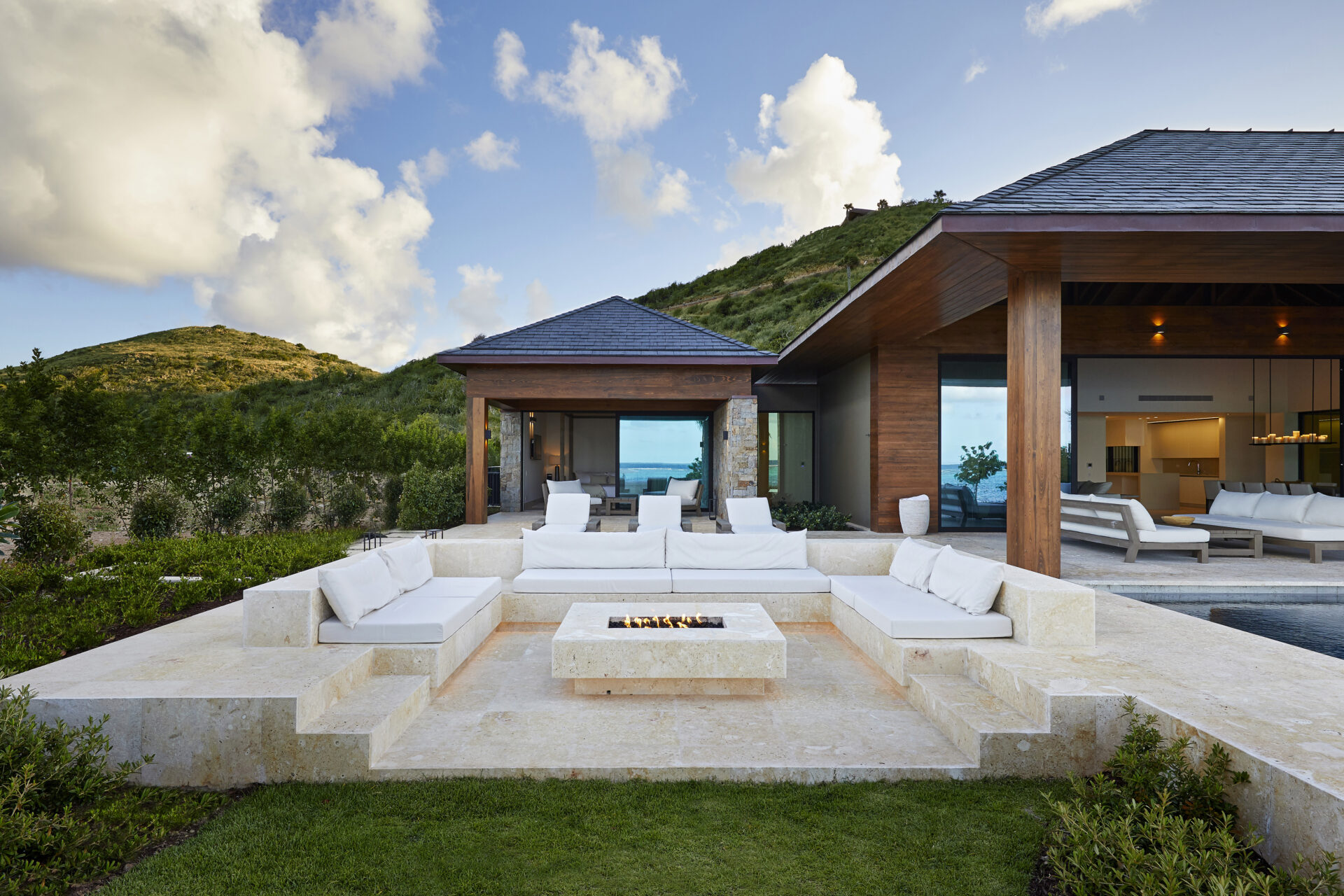 Modern luxury villa with outdoor seating area surrounding a fire pit, set against a backdrop of lush green hills. This beach house features large glass windows and sliding doors, leading to an indoor living area. The sky is partly cloudy.