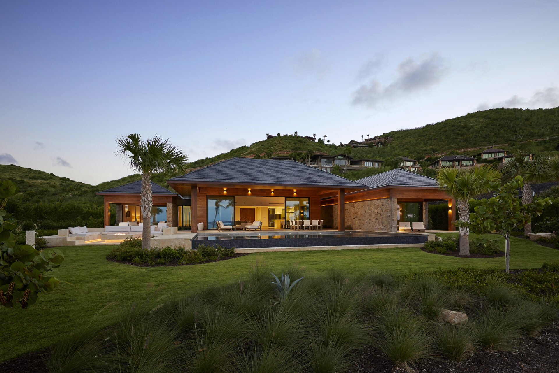 A luxurious single-story modern beach house with wooden accents and large glass doors sits surrounded by manicured lawns and tropical plants. The house features an open layout, a spacious patio, and a swimming pool, with lush green hills in the background under a blue sky.