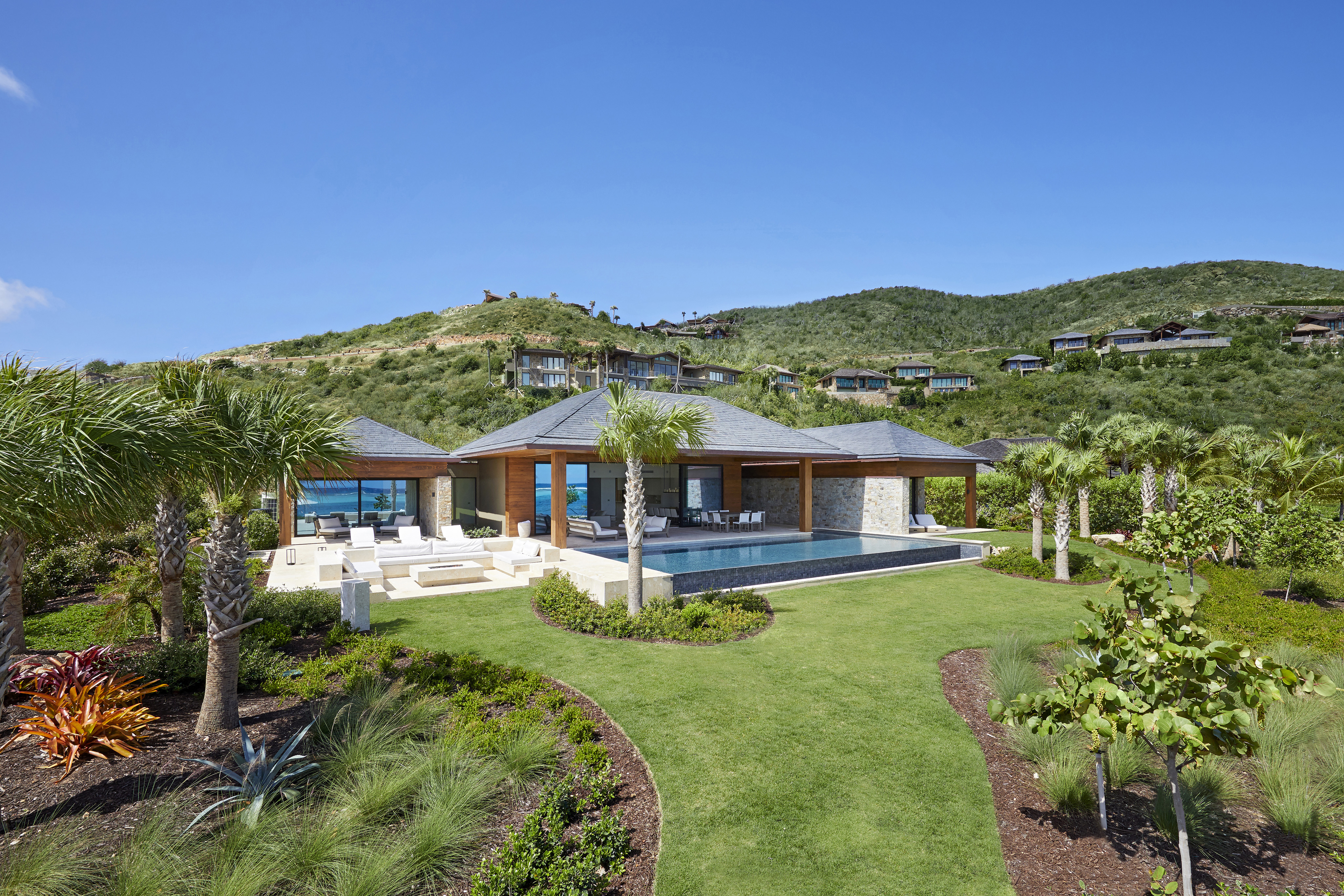 A modern, single-story house with a tiled roof and wooden accents is nestled among lush green hills near the beach. The home features a spacious outdoor patio, an infinity pool, and landscaped gardens with palm trees and colorful plants. The sky is clear and blue.