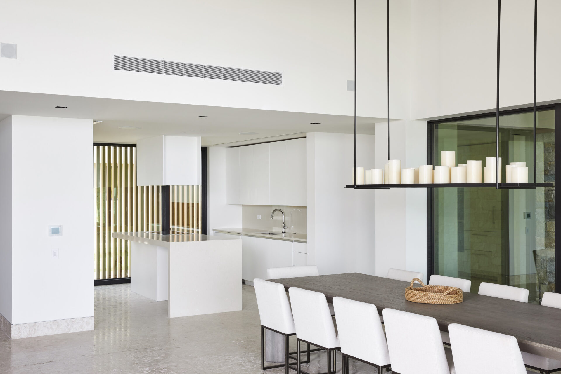 A modern kitchen and dining area with white walls and furniture perfect for a beach house. The kitchen boasts a large island with a white countertop and appliances, while the dining area features a long table with white chairs and a unique chandelier with white candles hanging above.